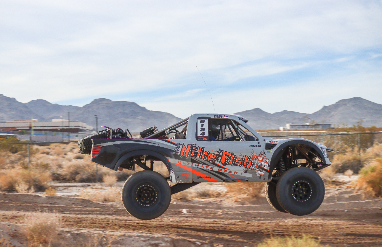 A racing dune buggy with large off-road tires jumping over a dirt track in a desert landscape with mountains in the background.