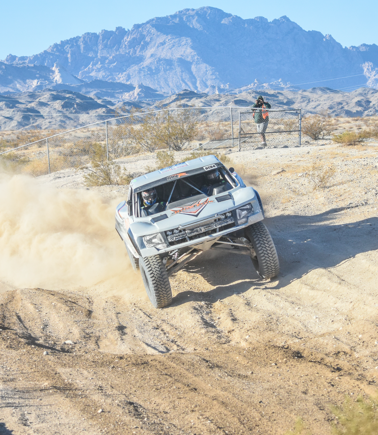 An off-road race car driving through a desert landscape, kicking up dust, with mountains in the background and a person watching from behind a fence.