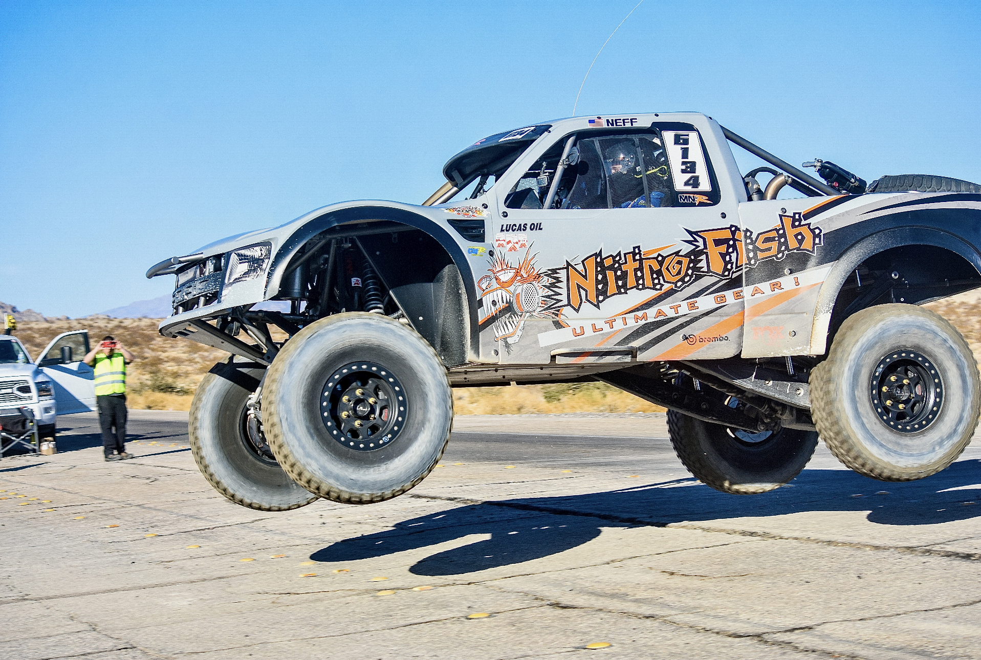 An off-road race truck with large tires, racing decals, and the words 'Nitro Fist' on the side, airborne during a race in a desert environment with a blue sky.