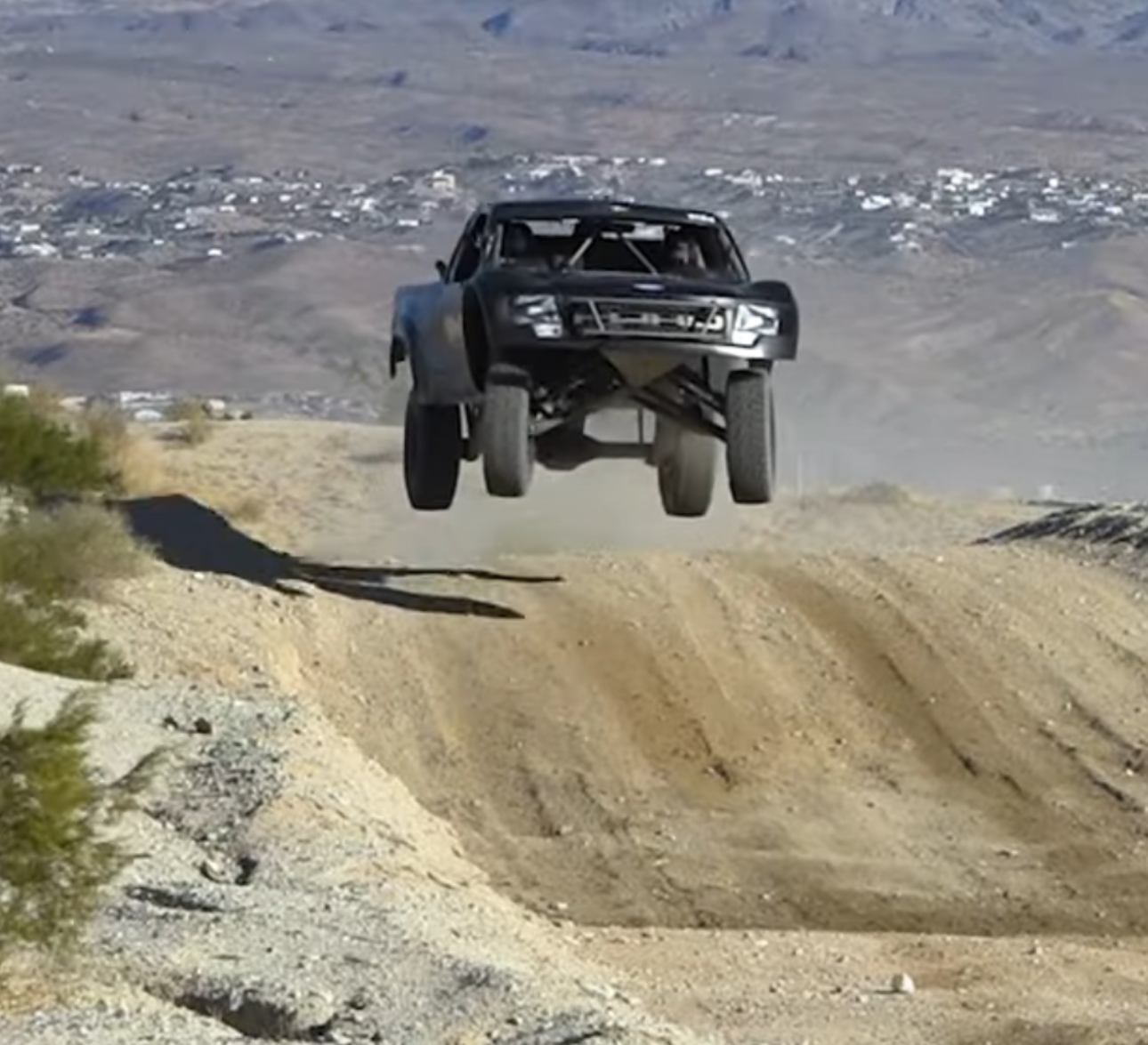A black off-road racing vehicle mid-air over a dirt track with mountains in the background.