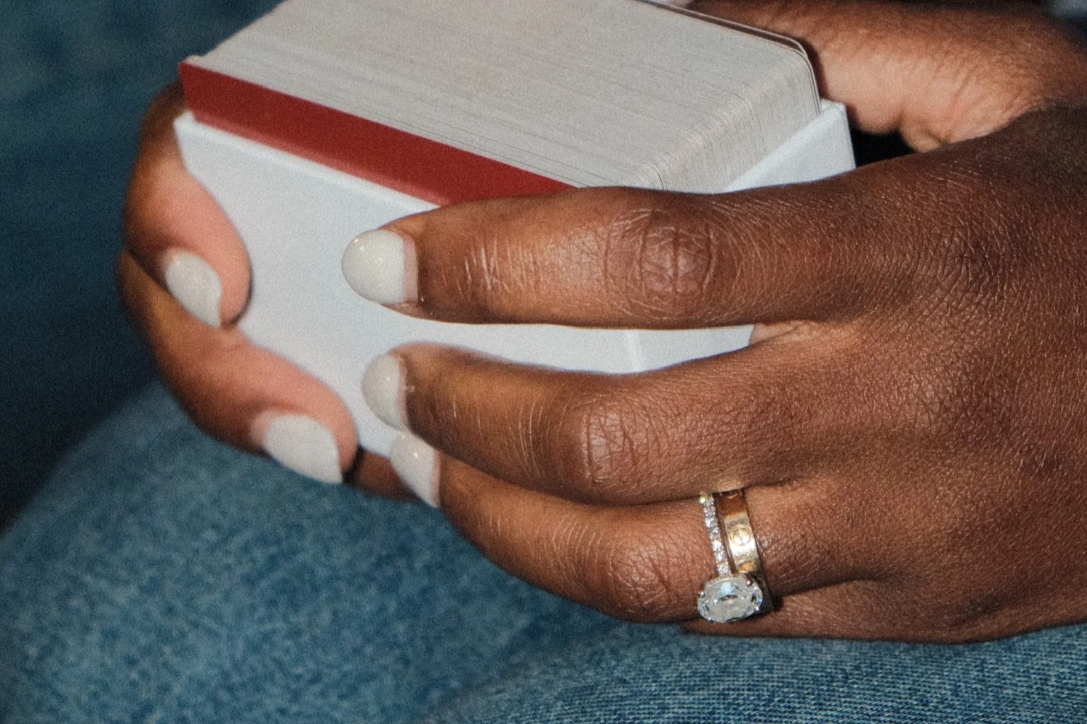 Close-up of a woman's hand holding a deck of playing cards. The woman has a gold ring with a large gemstone and smaller stones on her finger, bun ring, and painted white nails. The background includes denim fabric.