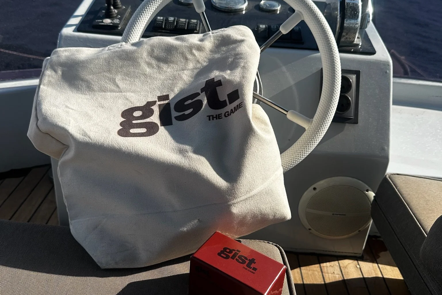 A boat's helm with a white steering wheel, a cloth bag labeled 'gist. THE GAME', and a red box also labeled 'gist. THE GAME' placed on a beige boat seat; the background shows part of the boat's interior and water outside.