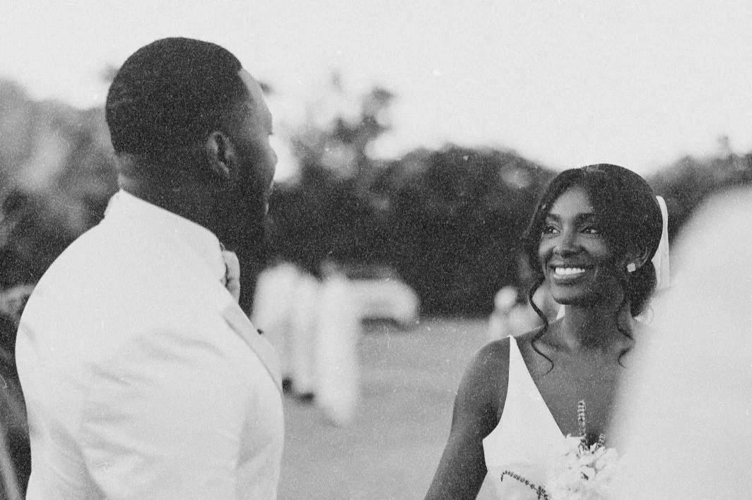 A black and white photograph of a bride and groom outdoors, facing each other with the bride smiling, during a wedding celebration.