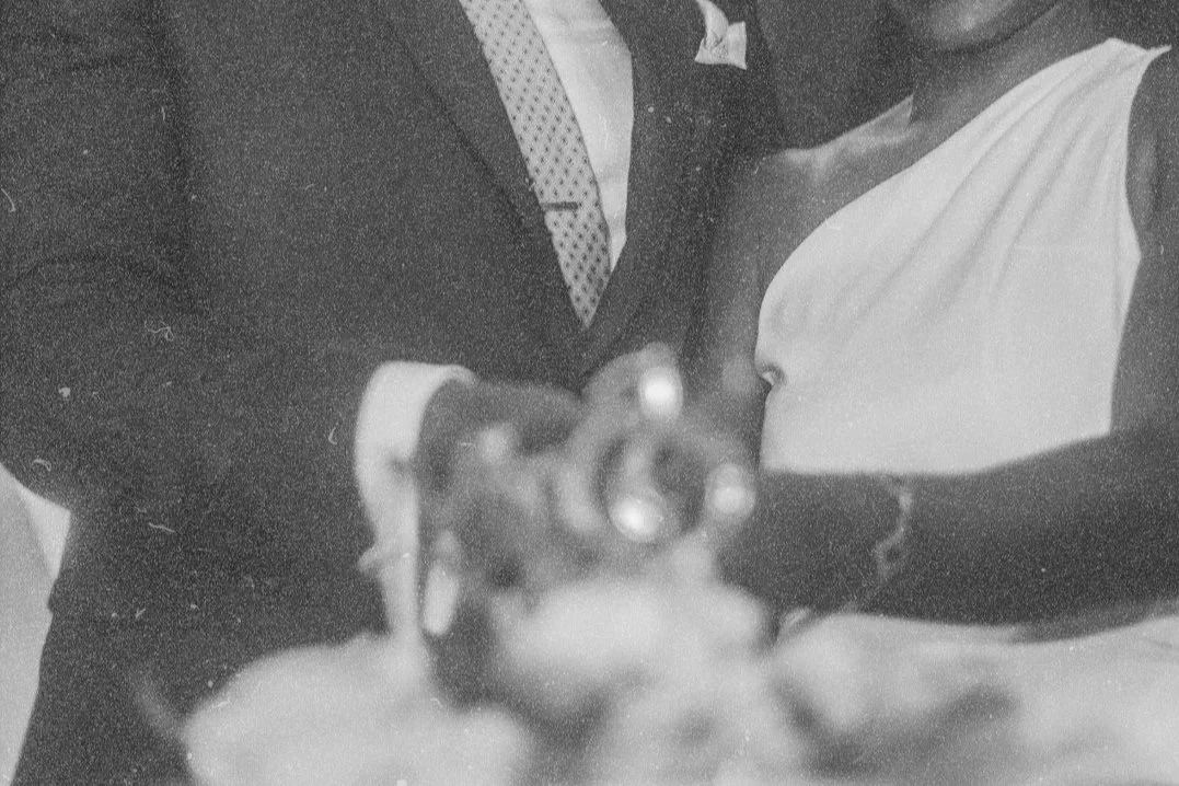 Two people, one in a suit and tie and the other in a white dress, are showing their rings towards the camera.