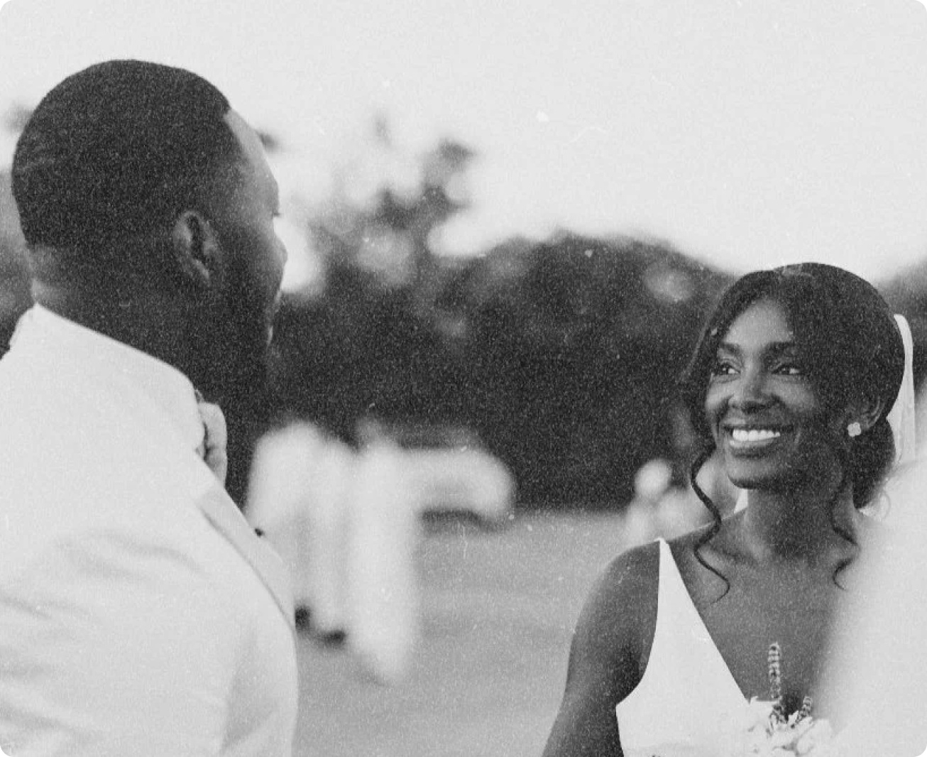 A black-and-white photo of a man and woman smiling at each other, outdoors, with blurred background trees, possibly during a wedding or formal event.