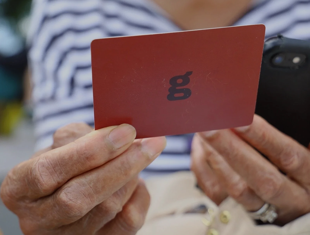 Close-up of a person holding a red card with the letter 'g' upside down, with another person holding a phone in the background.