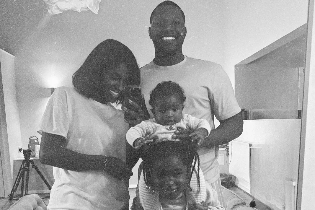 A family of four, including a woman, man, and two young children, taking a selfie in a mirror in a living room.