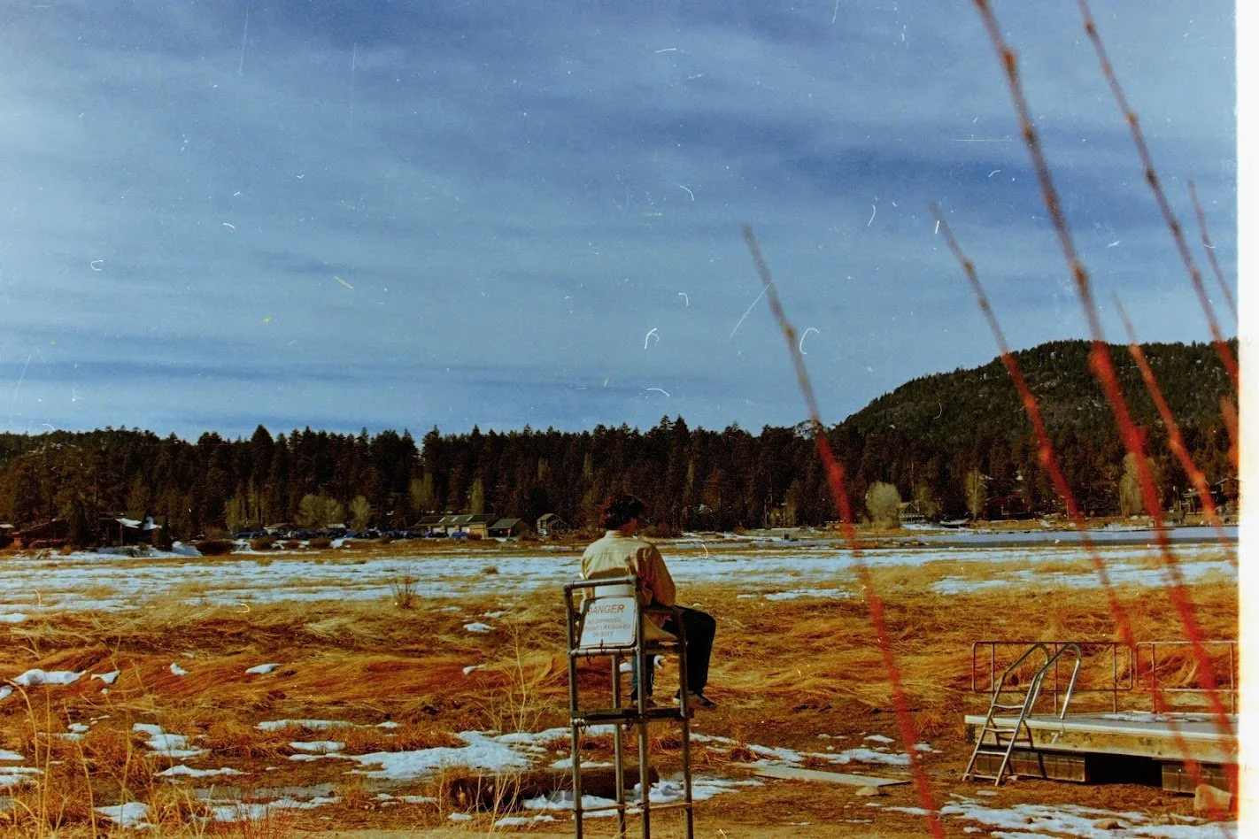 A person sitting on a lifeguard chair near a frozen lake, with houses and trees in the background, and mountains under a cloudy sky.