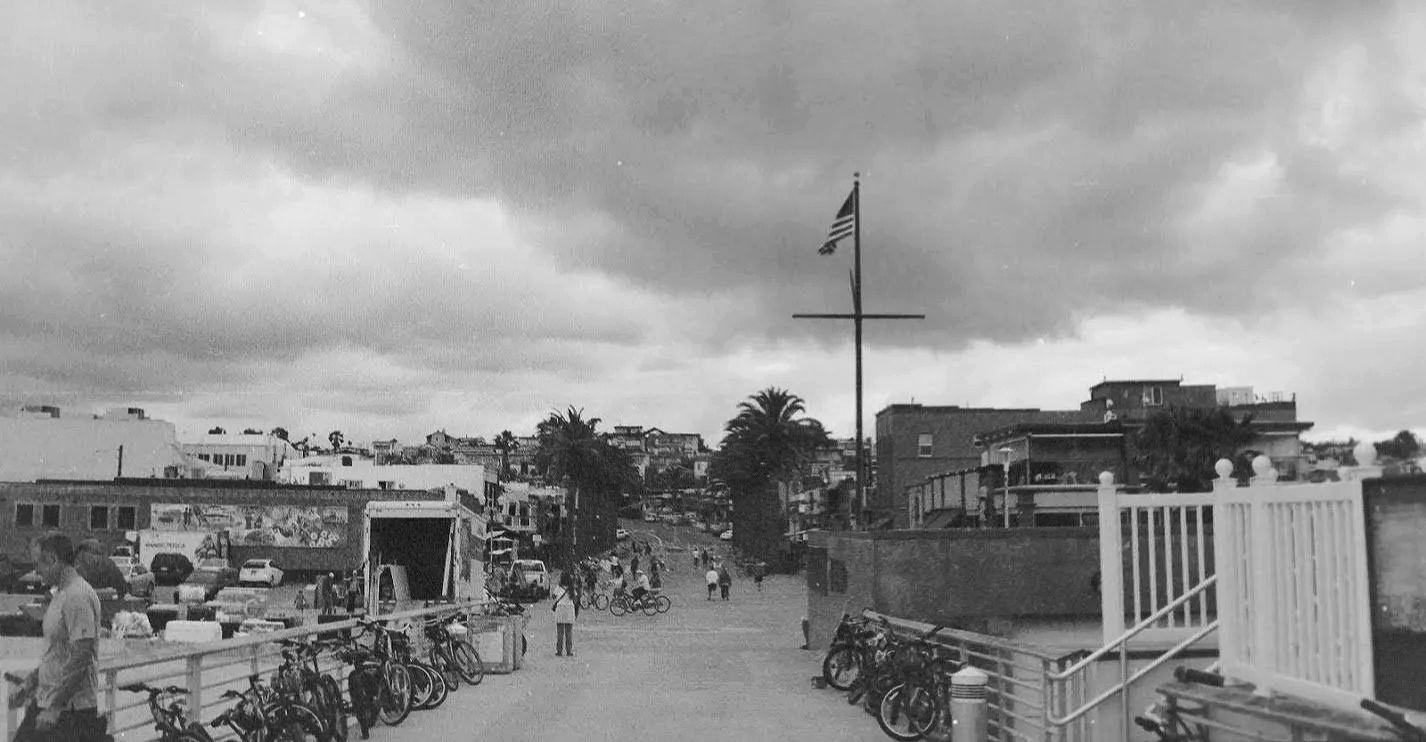 A black-and-white photo of a coastal town with a cloudy sky, palm trees, bicycles, cars, people walking, and an American flag on a flagpole.