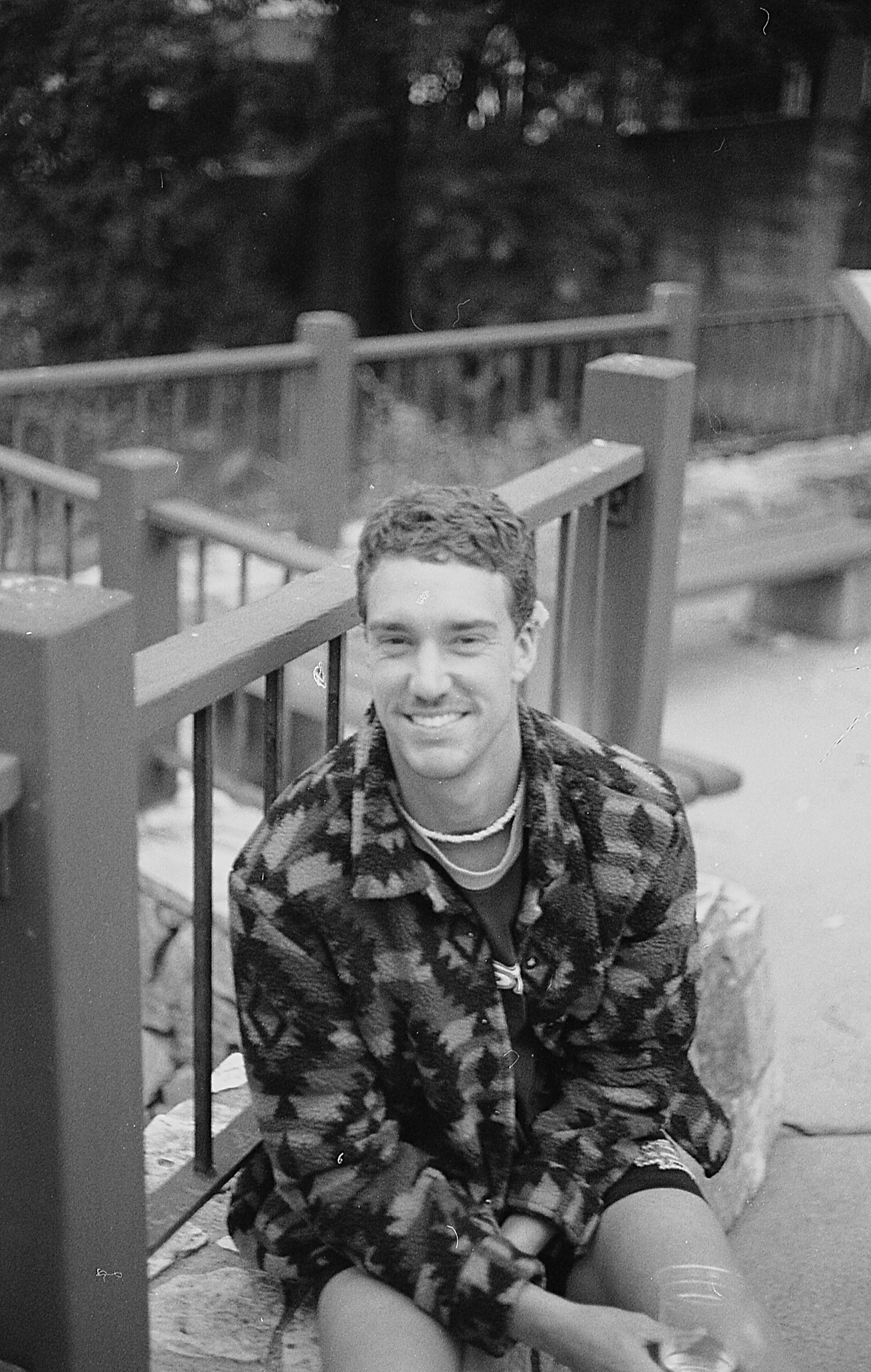A young man smiling, sitting outdoors near a wooden fence, wearing a camouflage jacket and shorts, with trees in the background in san diego county.