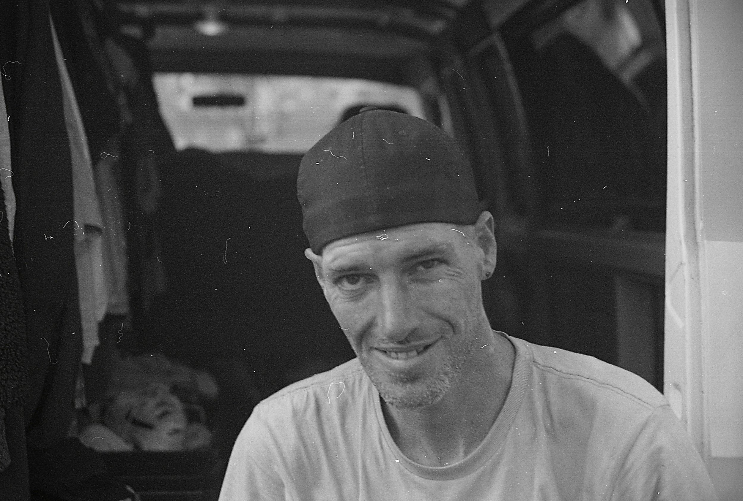 A black and white photo of a smiling man with a beard and mustache, wearing a dark beanie and a light-colored T-shirt, sitting inside a vehicle or similar enclosed space.