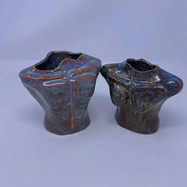 Two abstract ceramic vases with irregular shapes and dark, glossy glaze, placed on a plain surface against a light-colored background.