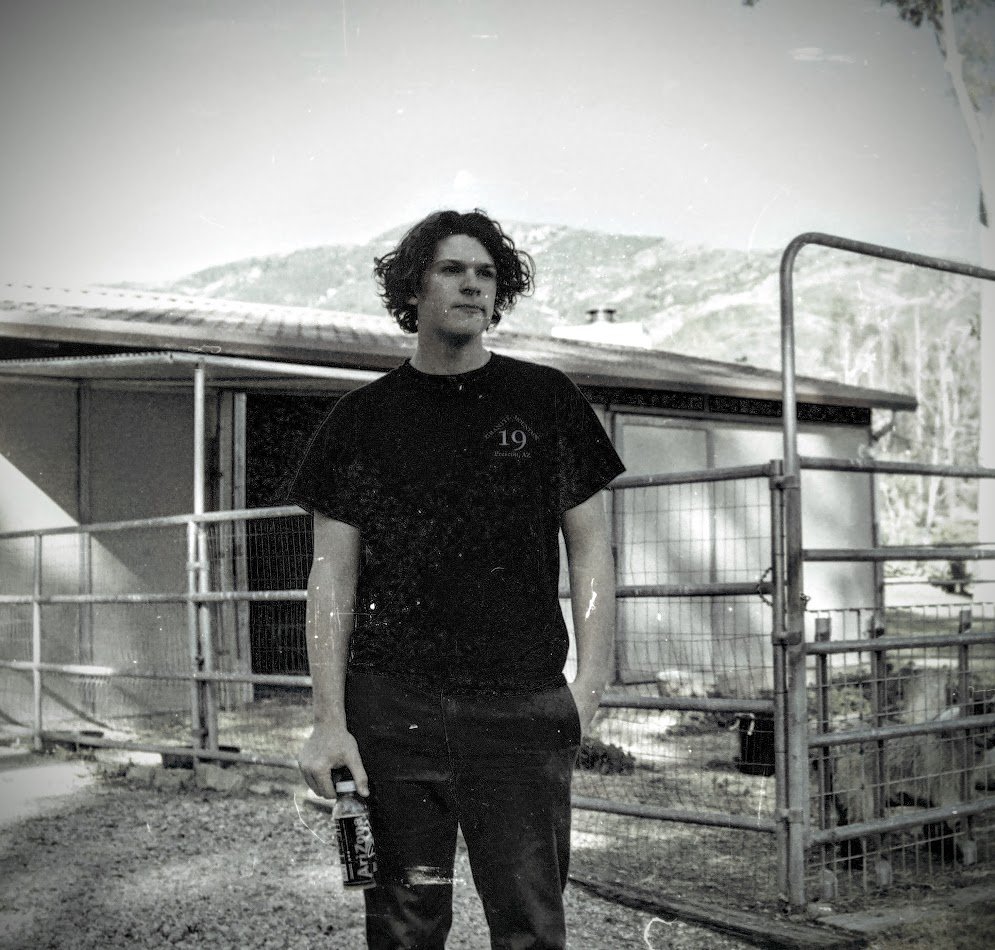 A young man with curly hair stands outdoors near a metal fence, holding a beverage bottle, with mountains and a building in the background in santa barbara.