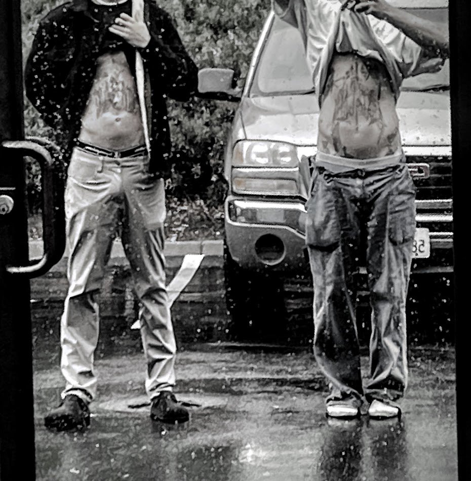 Two shirtless individuals taking mirror selfies outdoors on a rainy day, with a parked car in the background, wearing jackets or hoodies. One person has their hand on their chest, showing their torso.