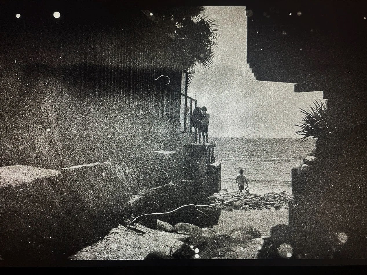 Black and white photo of two people embracing on a balcony overlooking a beach, with a child walking toward the water and ocean waves in the background, framed by plants and rocks.