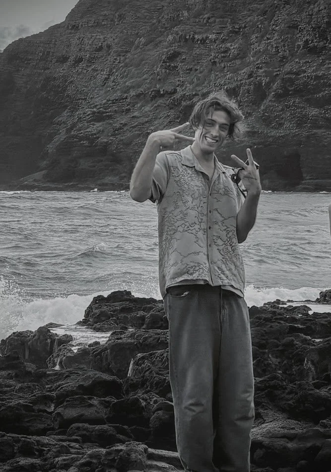 A young man posing on a rocky shoreline with ocean waves and cliffs in the background, smiling and making peace signs with both hands in hawaii.