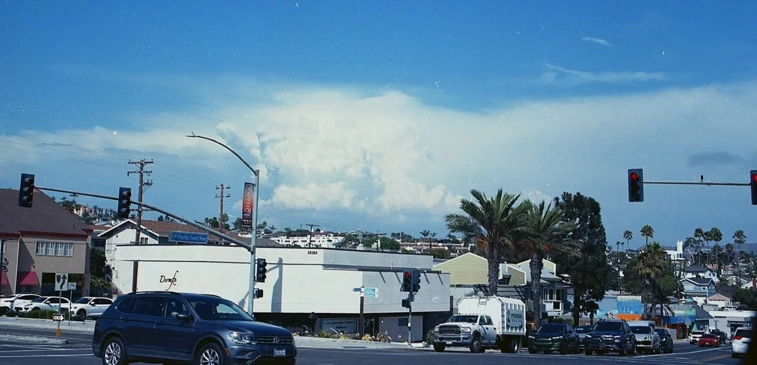 City street scene with cars, traffic lights, palm trees, and buildings under a cloudy sky.