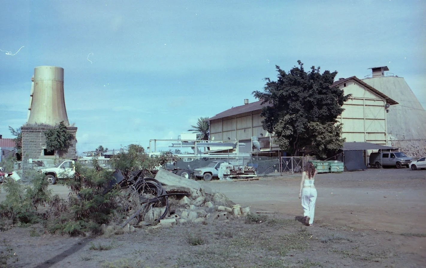 A woman in white walking near a junkyard with plants, old cars, and industrial structures in the background under a blue sky.
