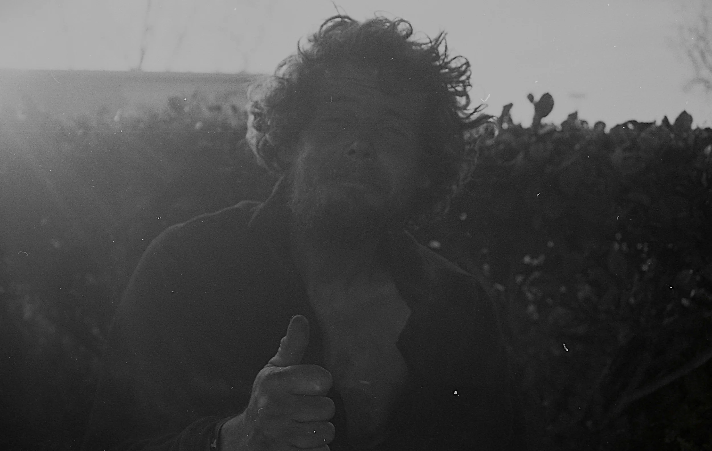 A man with curly hair and a beard giving a thumbs-up gesture in front of bushes or plants, in black and white.