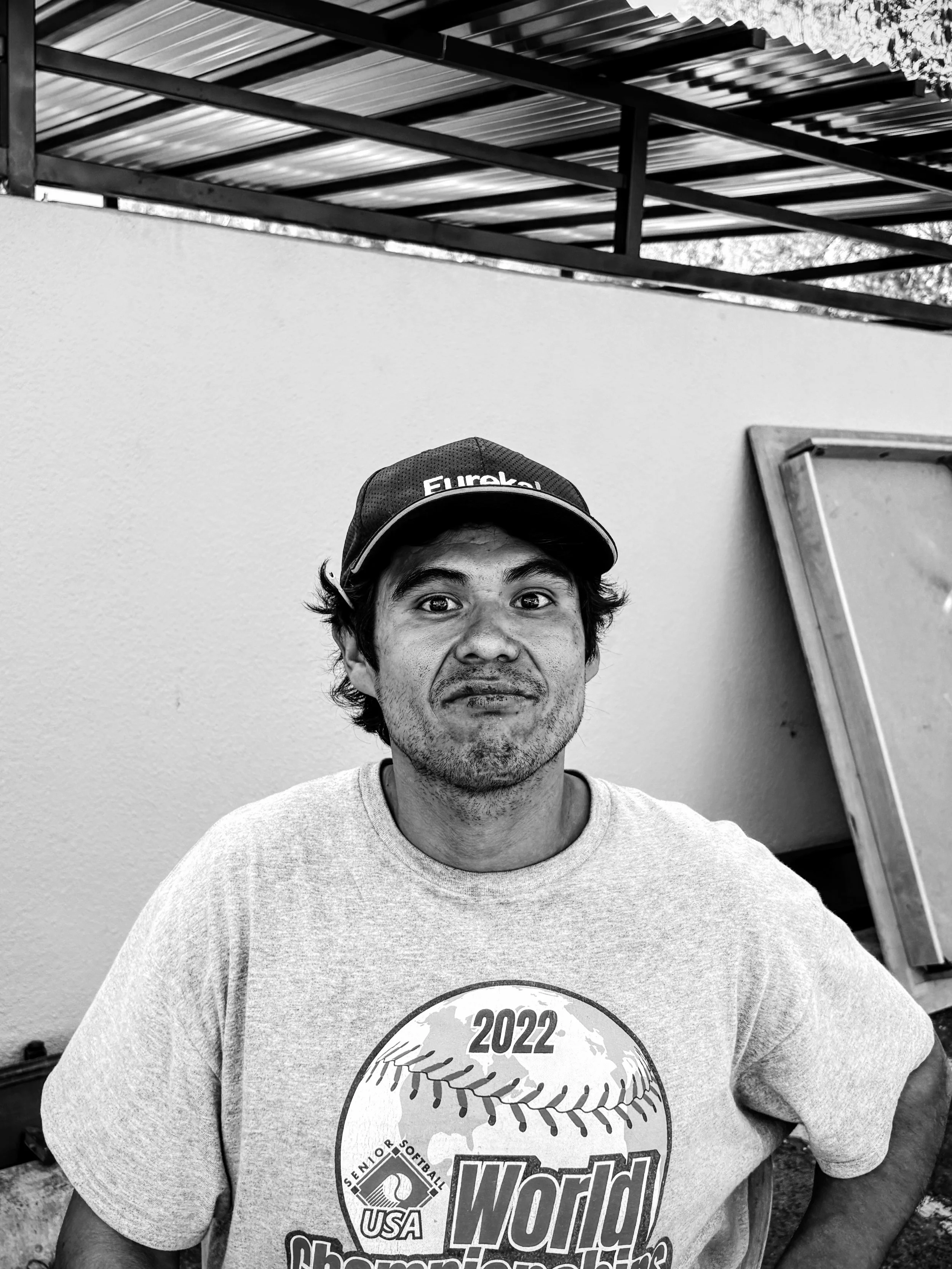 A man wearing a baseball cap and a T-shirt stands outdoors in front of a plain wall and a wooden frame, smiling slightly, in black and white.