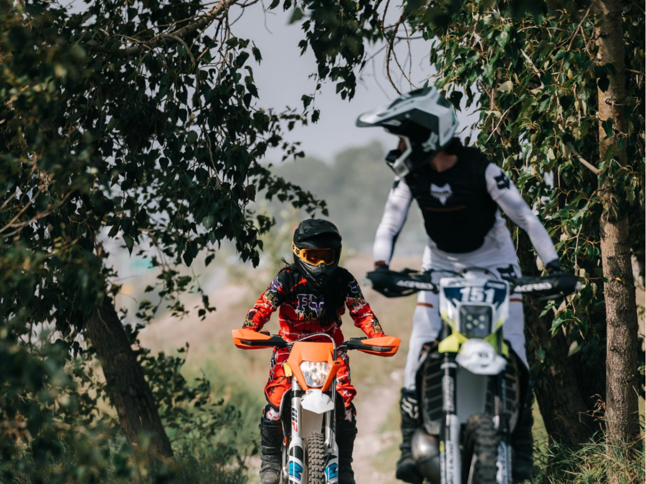 Two motocross riders, a child and an adult, riding dirt bikes on a trail through a wooded area, both wearing protective gear and helmets.