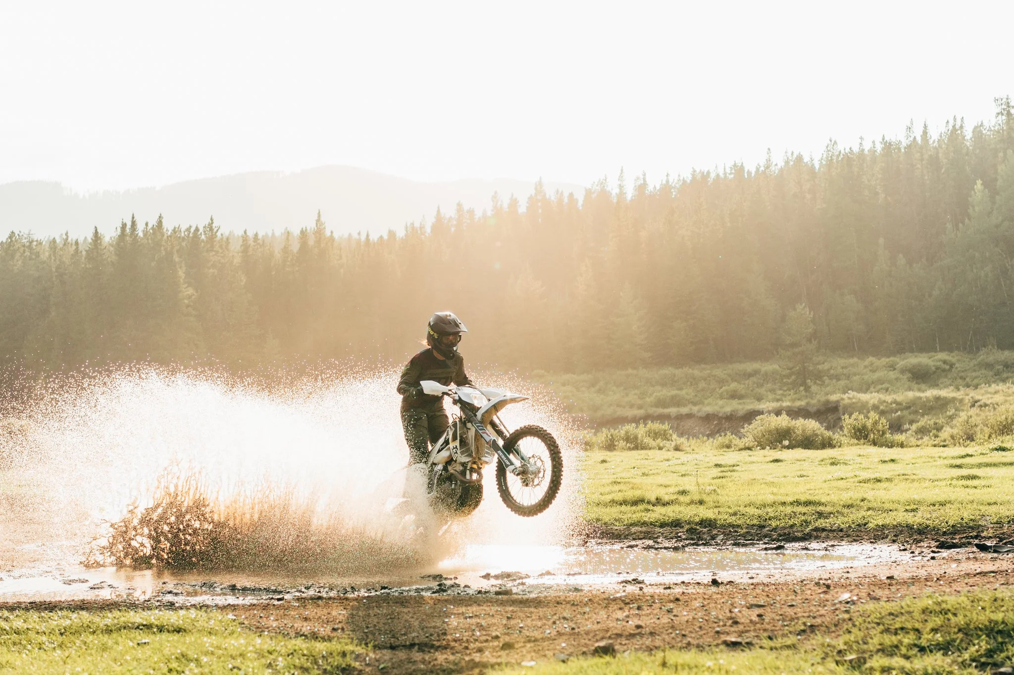 Person in black protective gear riding a dirt bike through water across a grassy area with trees and mountain in the background at sunset.