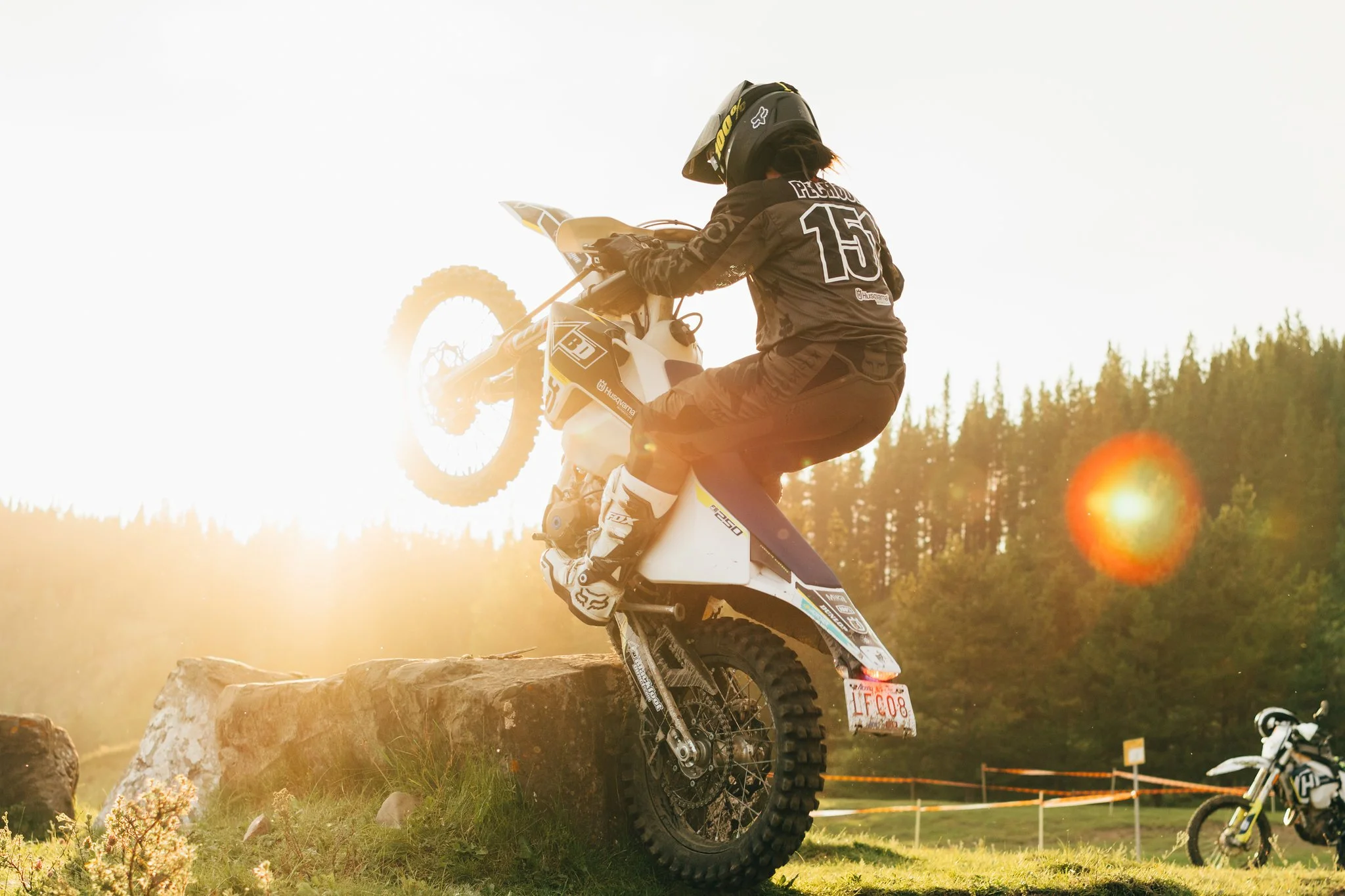 Motorcycle rider performing a jump off a rock during sunset, with a second motorcycle in the background and a forested area behind.