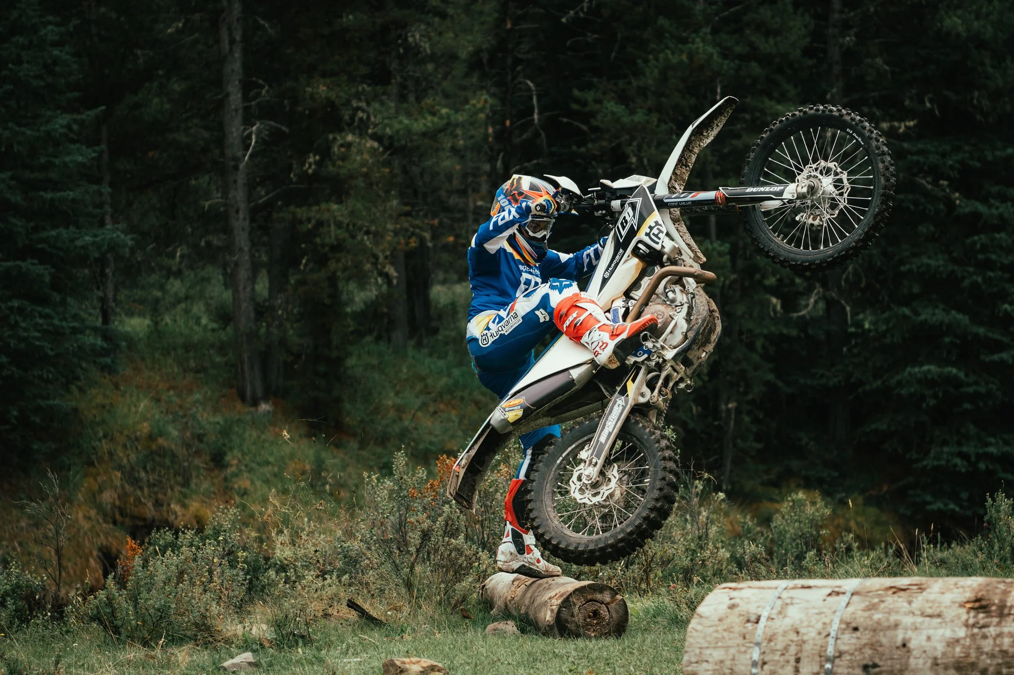 Motocross rider in blue gear balancing on a log with a dirt bike in a forest setting.