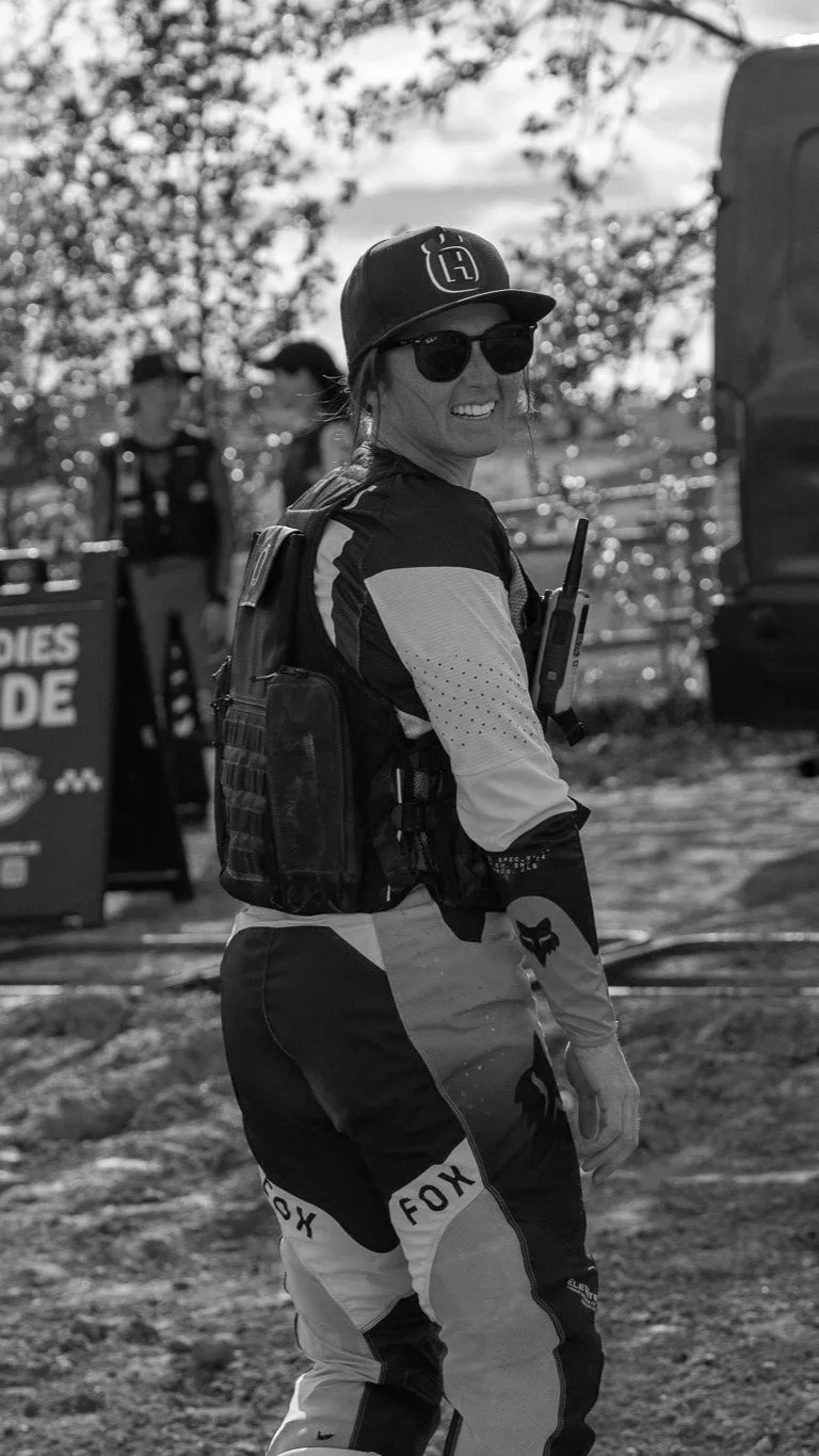 A woman wearing sunglasses, a cap, and motocross gear, smiling and standing outdoors during daytime with trees in the background.