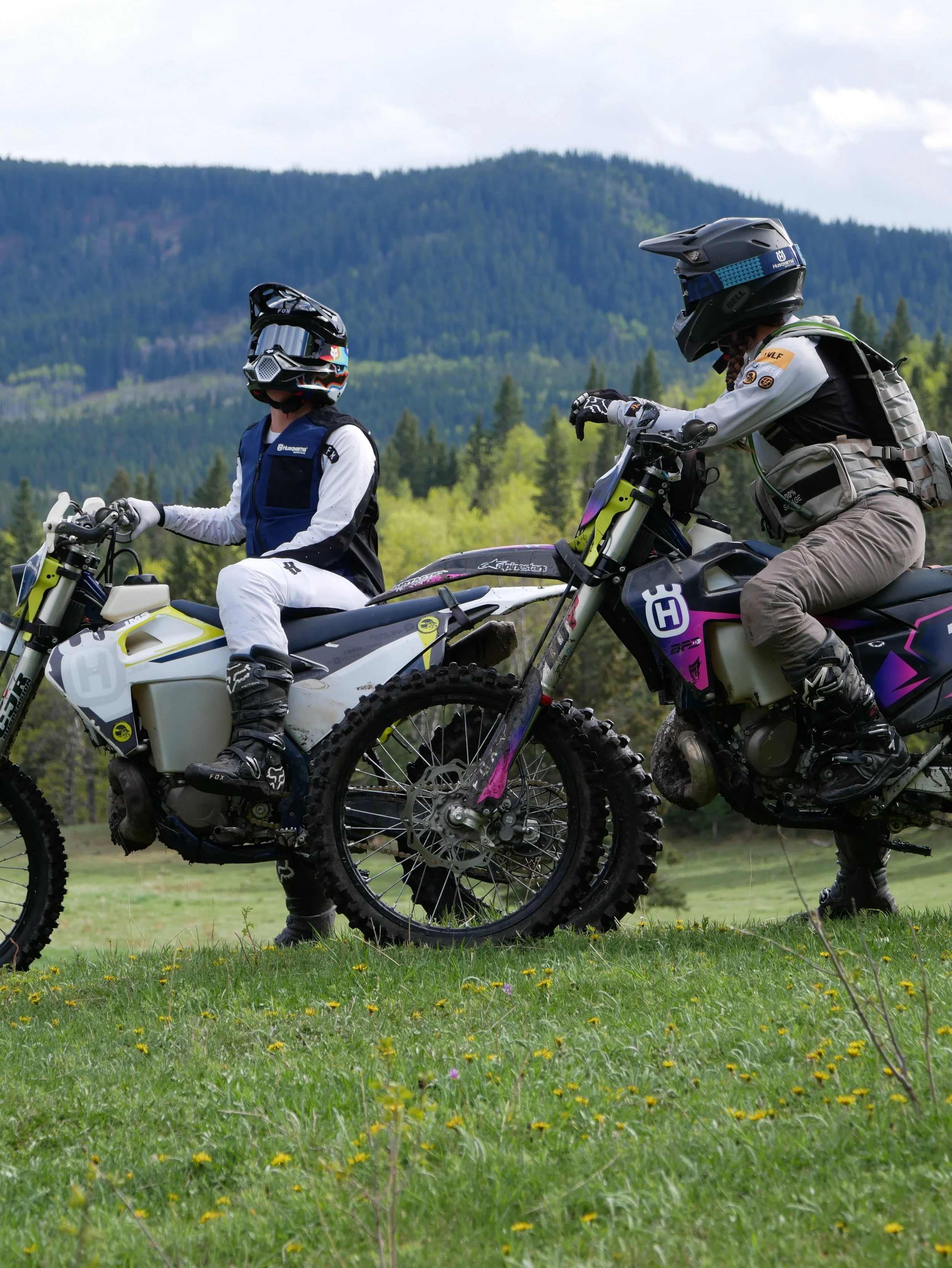 Two people riding off-road motorcycles in a grassy field with a mountain and forest backdrop.