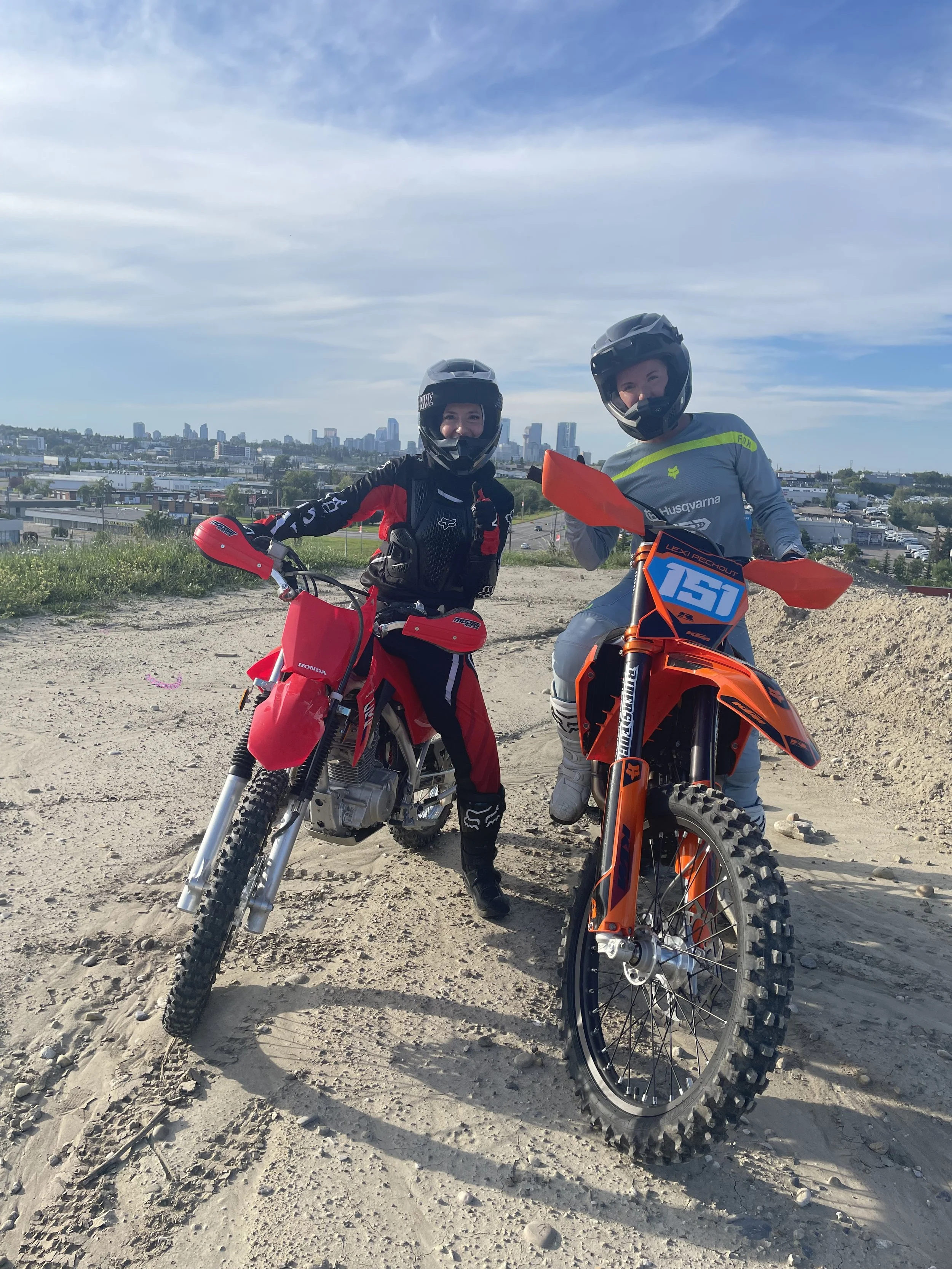 Two people in motocross gear and helmets standing with red dirt bikes on a dirt hill with city skyline in the background.