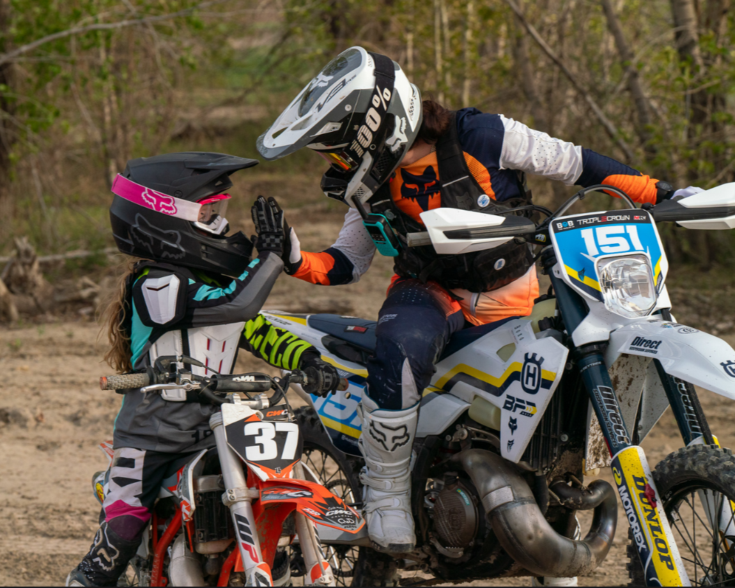 A woman and a girl in motocross gear giving each other a high five outdoors on dirt bikes.