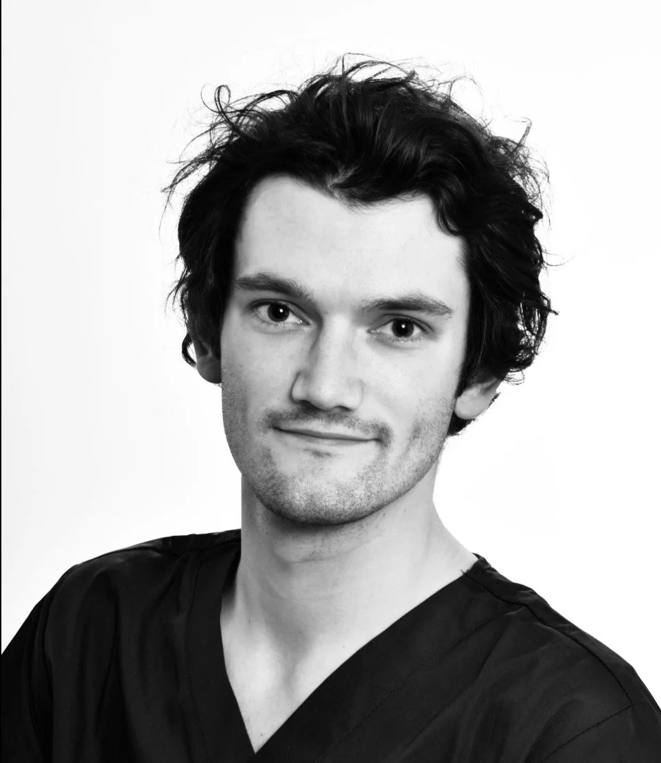 Black and white portrait of a young man with wavy dark hair, wearing a dark V-neck shirt, smiling slightly, against a plain background.