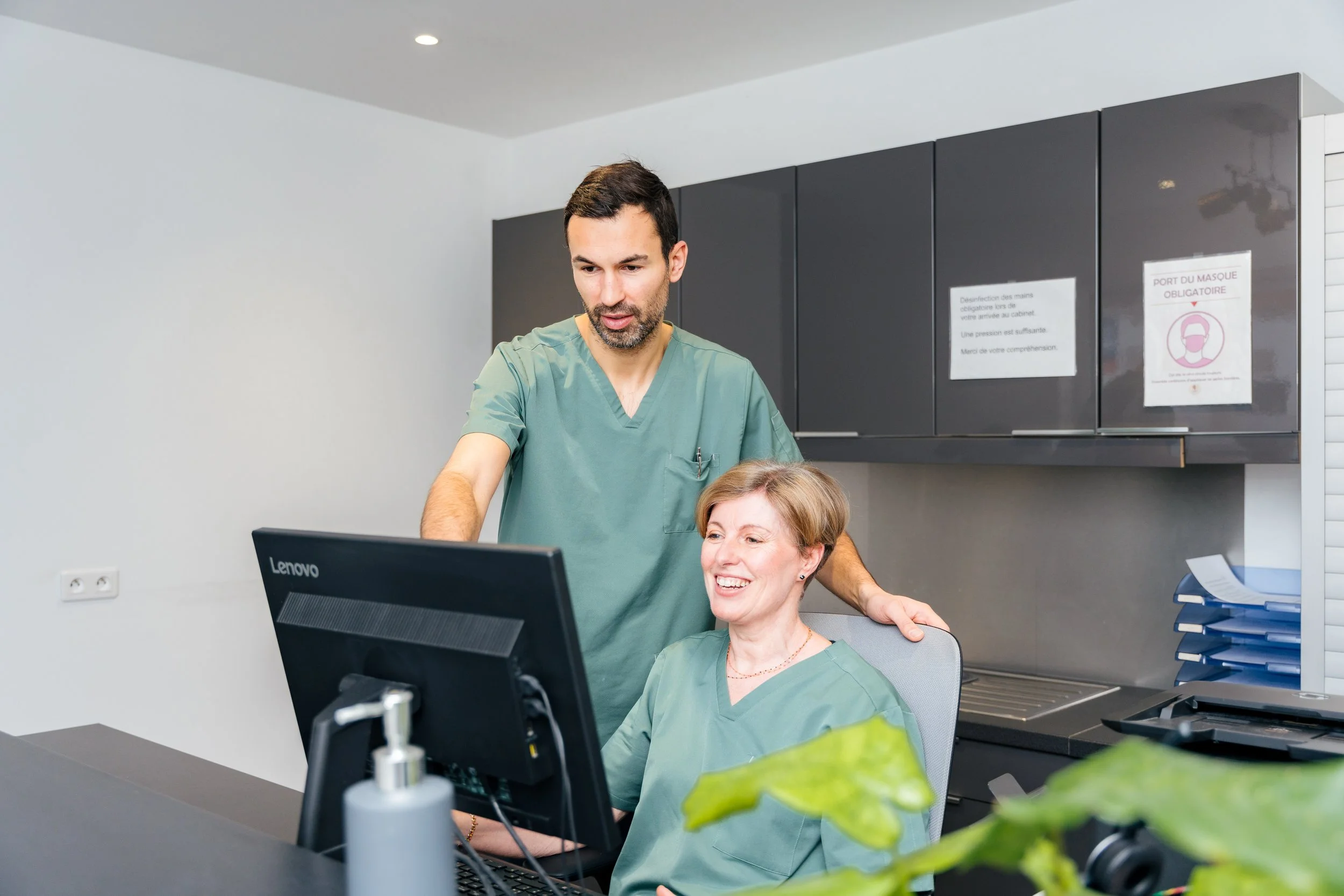 A healthcare professional in green scrubs assisting a smiling patient in green scrubs at a desk with a computer in a clinical setting. There are cabinets with signs on the wall and a plant in the foreground.