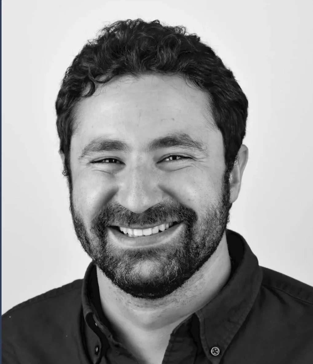 Black and white portrait of a smiling man with short, curly hair and a beard, wearing a collared shirt.