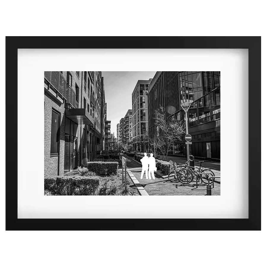 Black and white photograph of an urban street scene with modern buildings, bicycle racks, a streetlamp, and two people standing on the sidewalk.