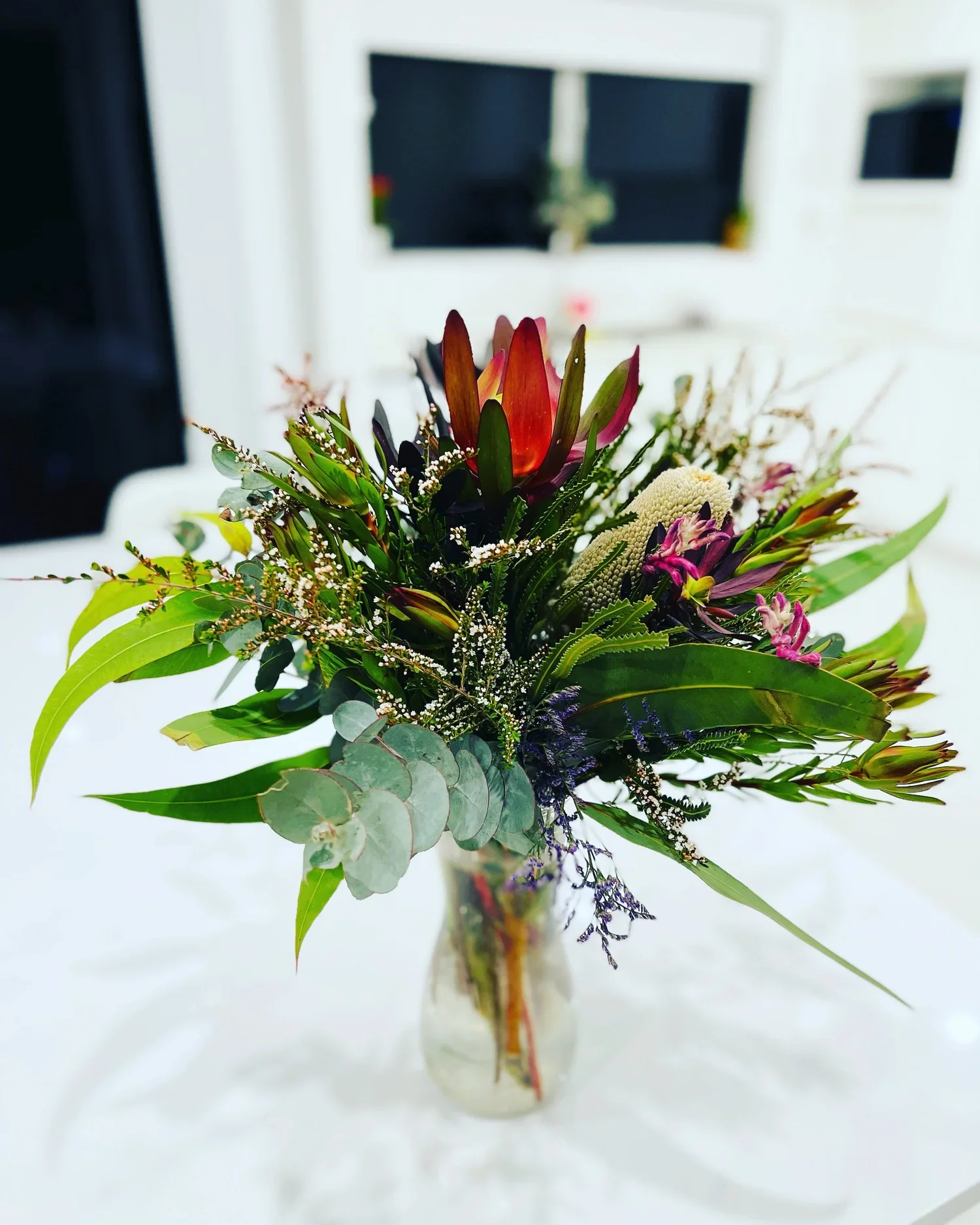 A colorful bouquet of flowers in a clear glass vase, featuring various types of greenery and blooms, on a white surface with a blurred background of a television or monitor.