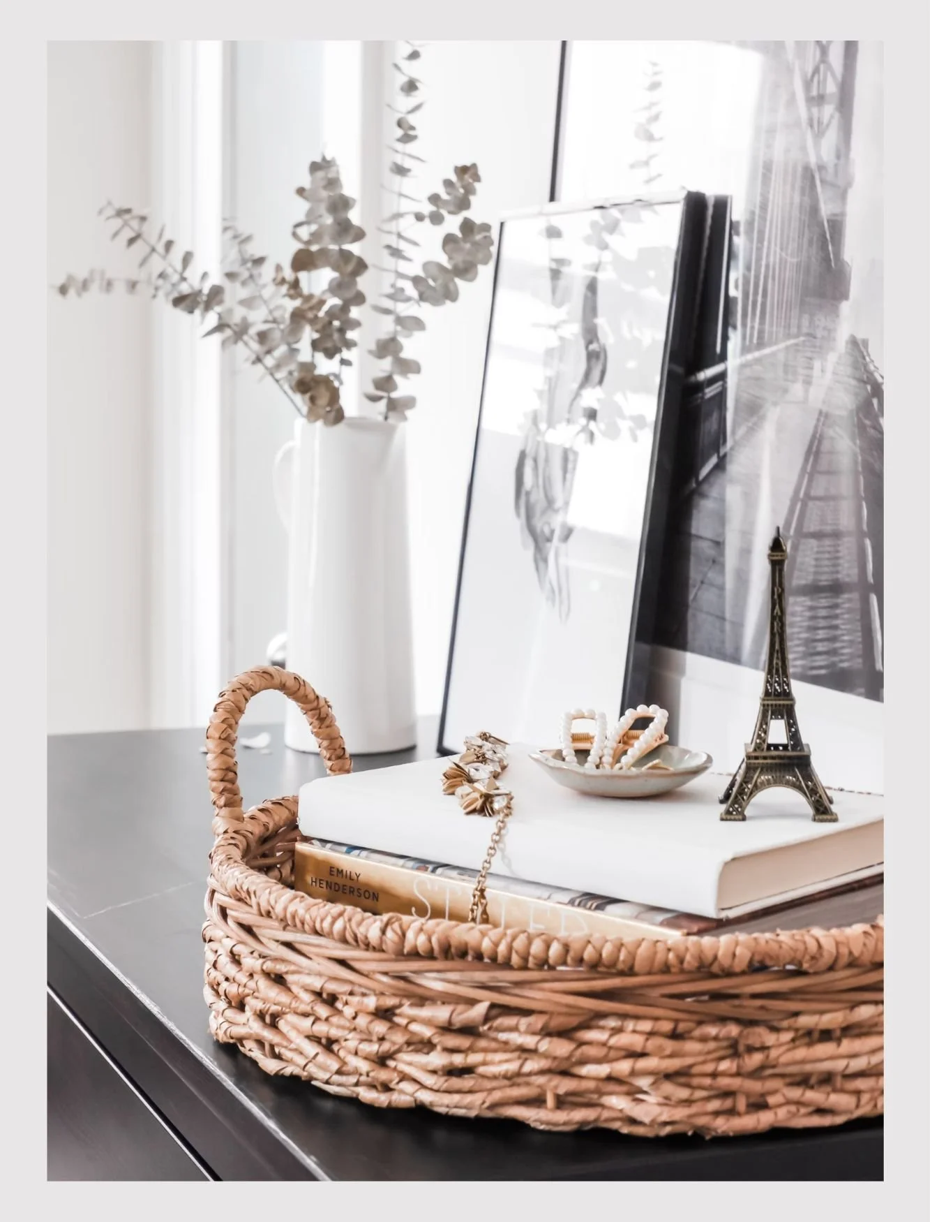Decorative tabletop arrangement with a wicker basket holding books, jewelry, and a mini Eiffel Tower replica, with framed artwork and dried eucalyptus in a white vase in the background.
