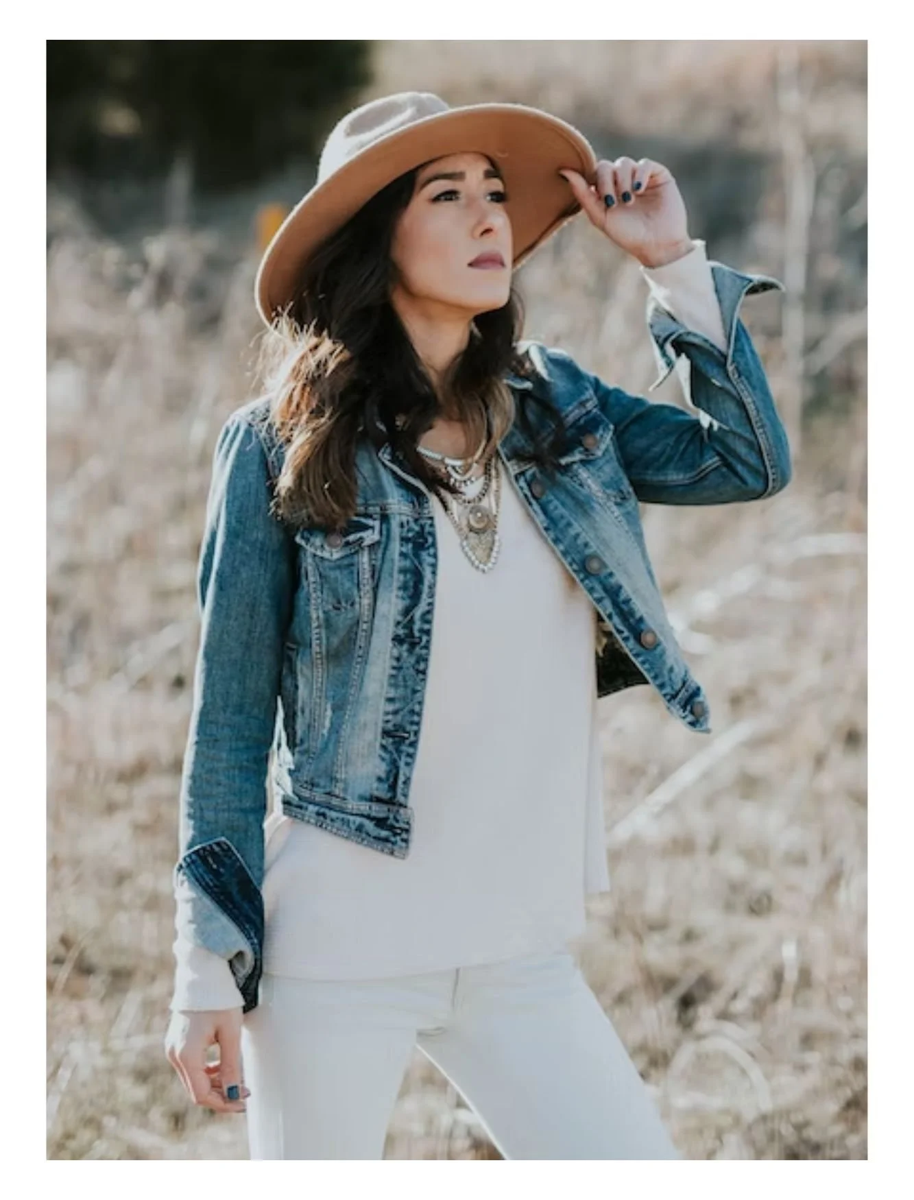 Woman in a denim jacket and white outfit wearing a wide-brim hat outdoors, hand adjusting her hat, with a background of dry grass.