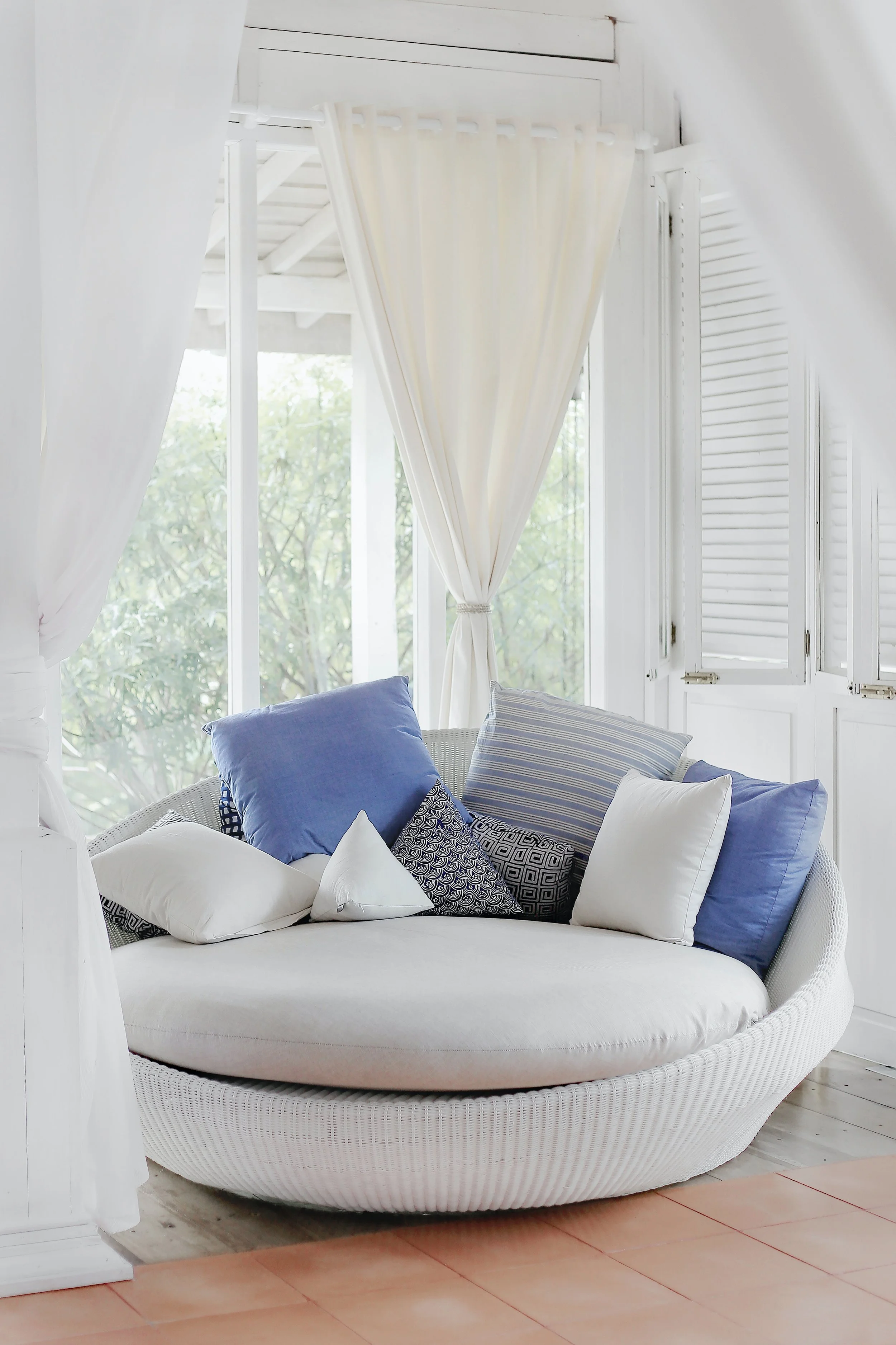 A round white wicker daybed with multiple blue and white pillows, situated in a bright room with white curtains and white shutters, overlooking trees outside.