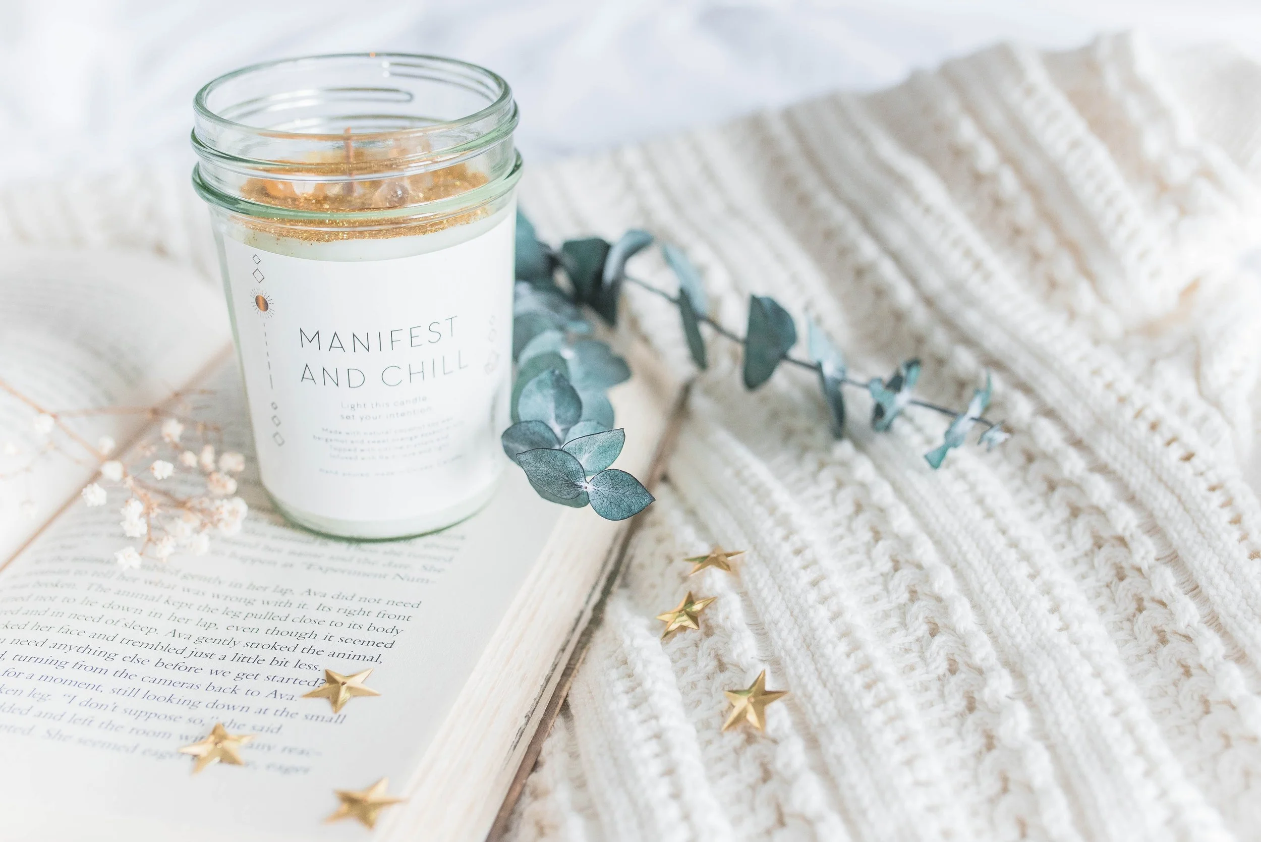 A white candle labeled 'Manifest and Chill' in a glass jar, placed on a book with in a cozy setting featuring a white knitted blanket, gold star confetti, eucalyptus leaves, and dried flowers.