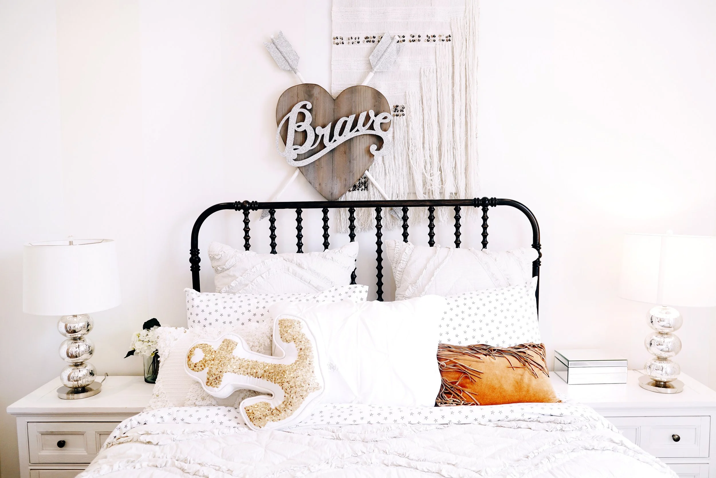 A bedroom featuring a black metal bed with white bedding and decorative pillows. Above the bed, there is a wall hanging with a wooden heart, arrows, and the word 'Brave' in cursive. On either side of the bed, there are white nightstands with silver lamps, a small flower arrangement, and a decorative box.