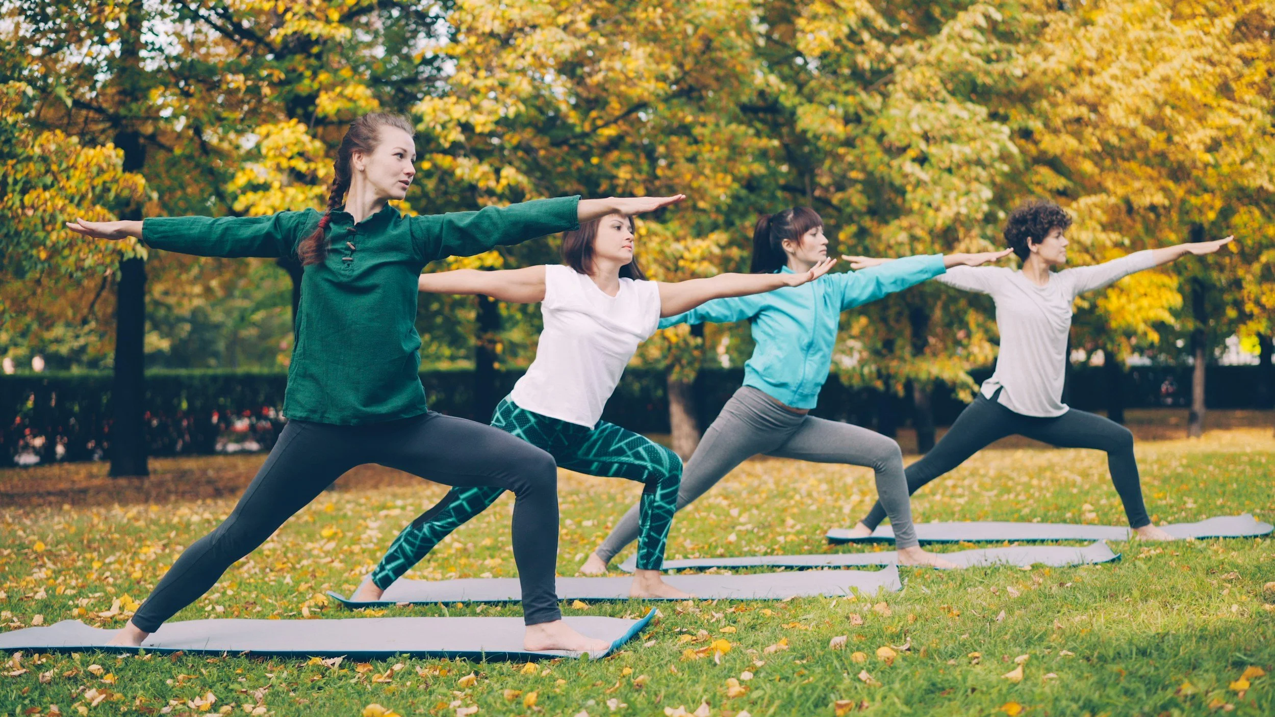 Four women practicing yoga outdoors on mats in a park with autumn leaves, doing warrior pose with arms extended.