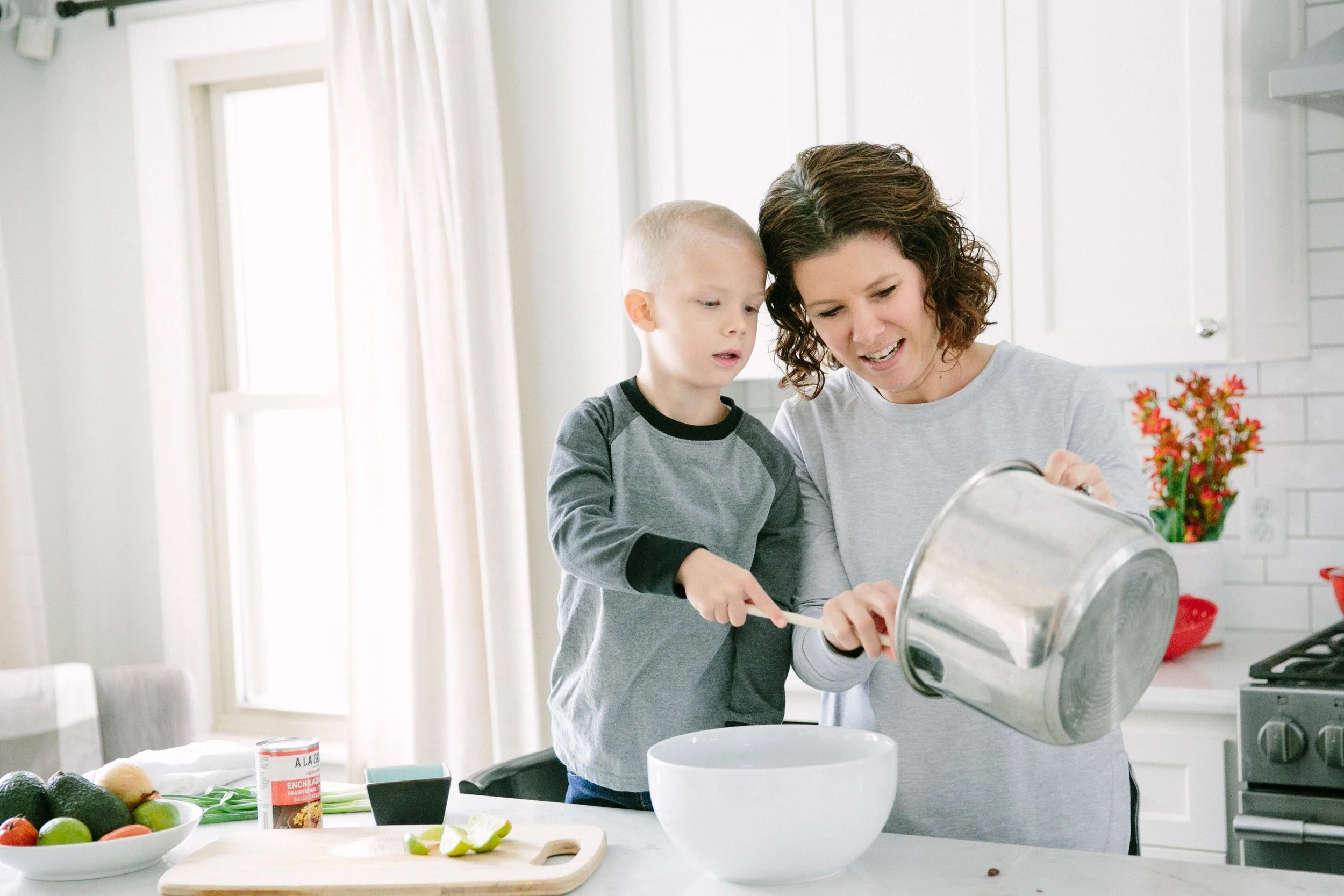 A woman and a young boy cooking together in a bright kitchen, pouring ingredients into a bowl.