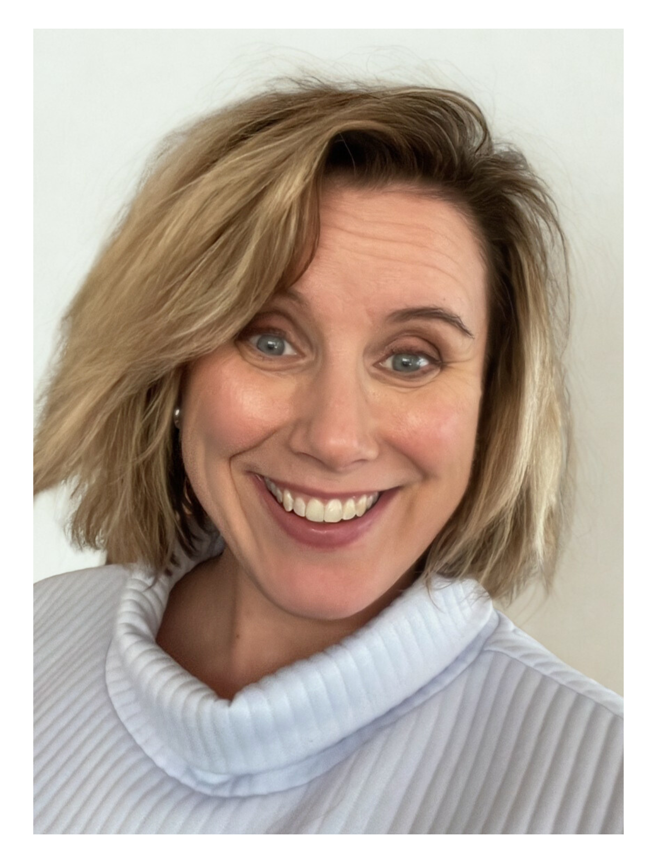Close-up photo of a woman smiling, with short, wavy blonde hair, wearing a white ribbed turtleneck sweater, against a plain light background.