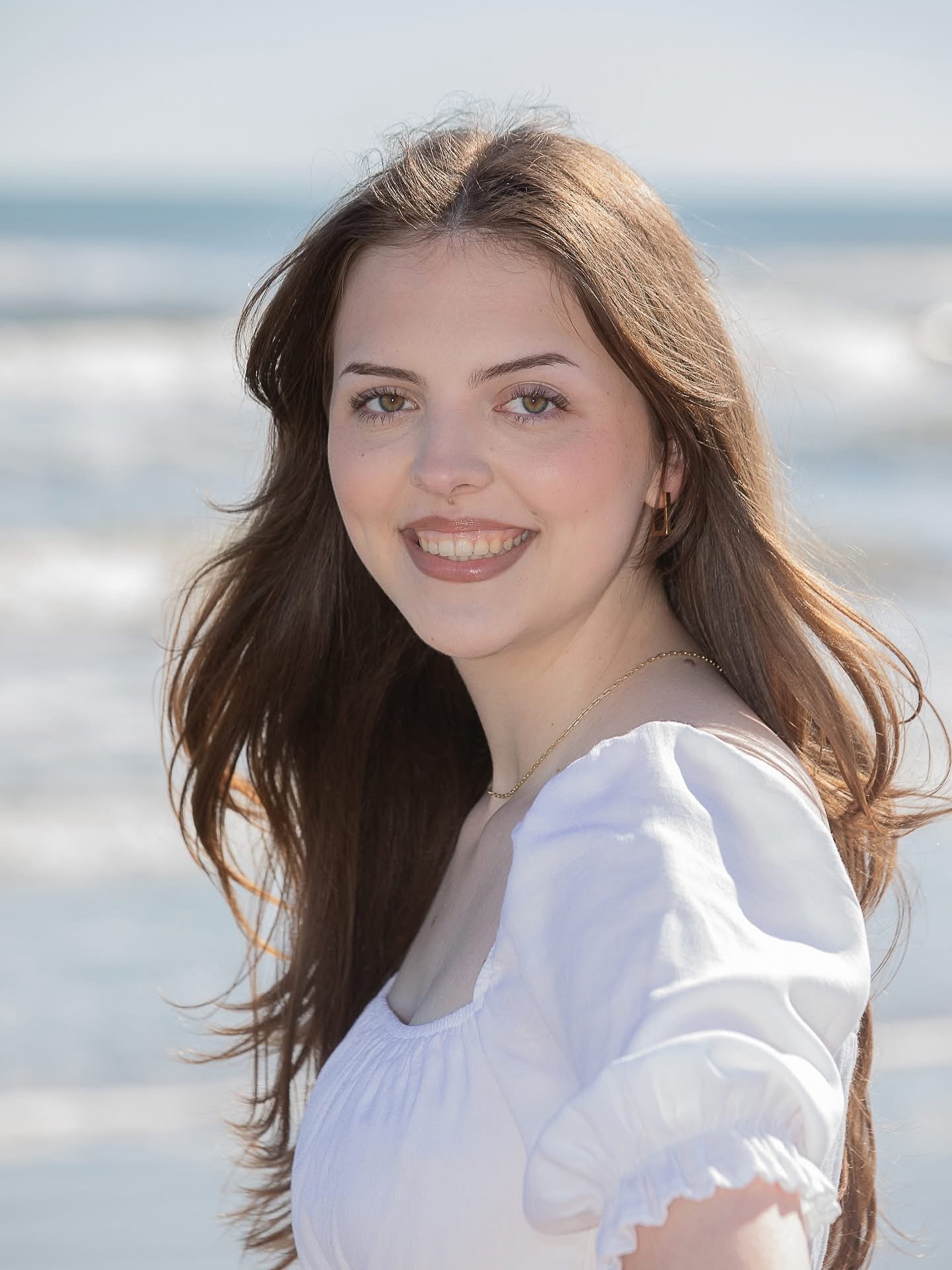 A young woman with long brown hair, green eyes, and light skin, smiling while standing on a beach with ocean waves in the background.
