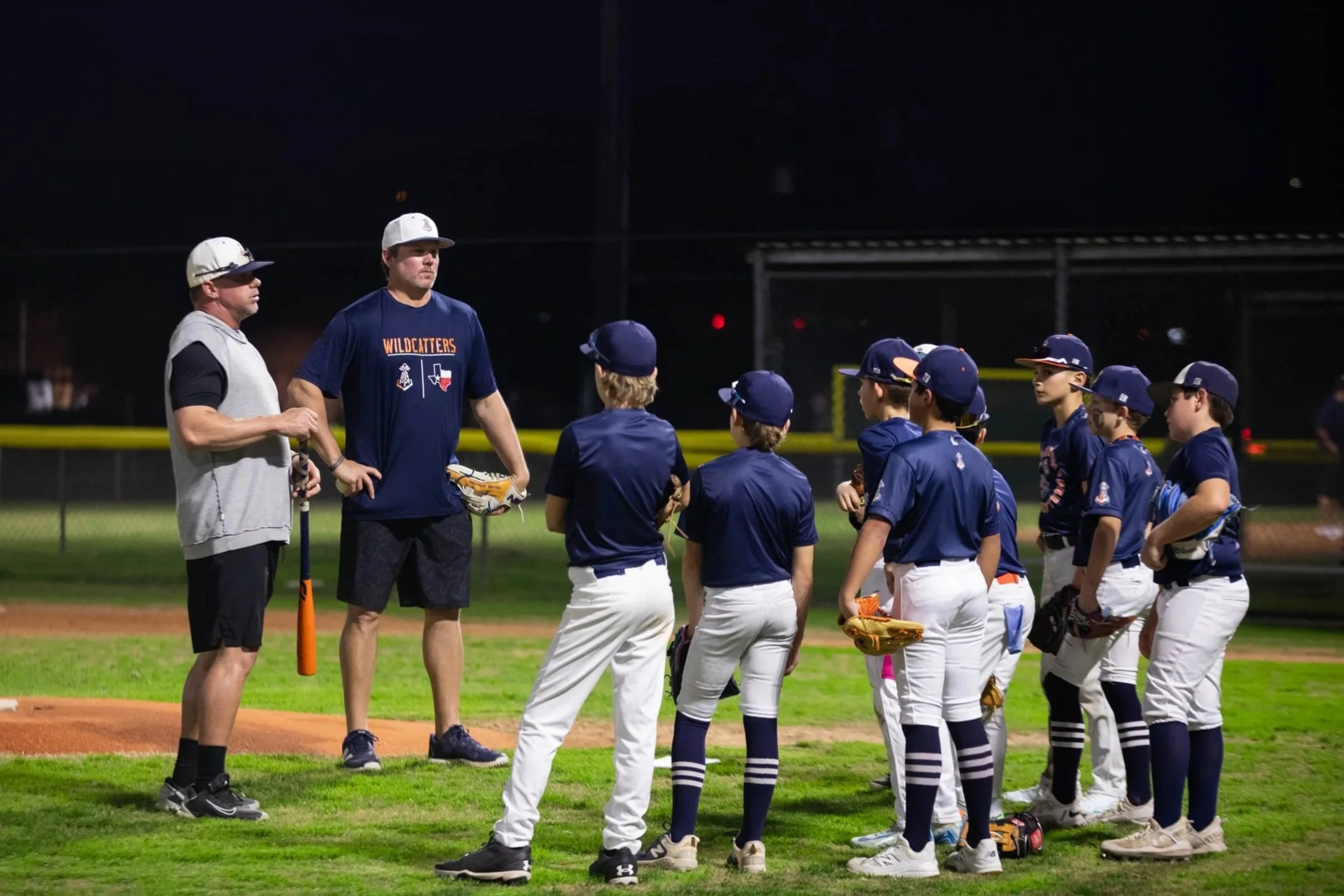 A youth baseball team listening to their coaches on a baseball field at night, with players in navy blue jerseys and white pants, standing near the pitcher's mound.
