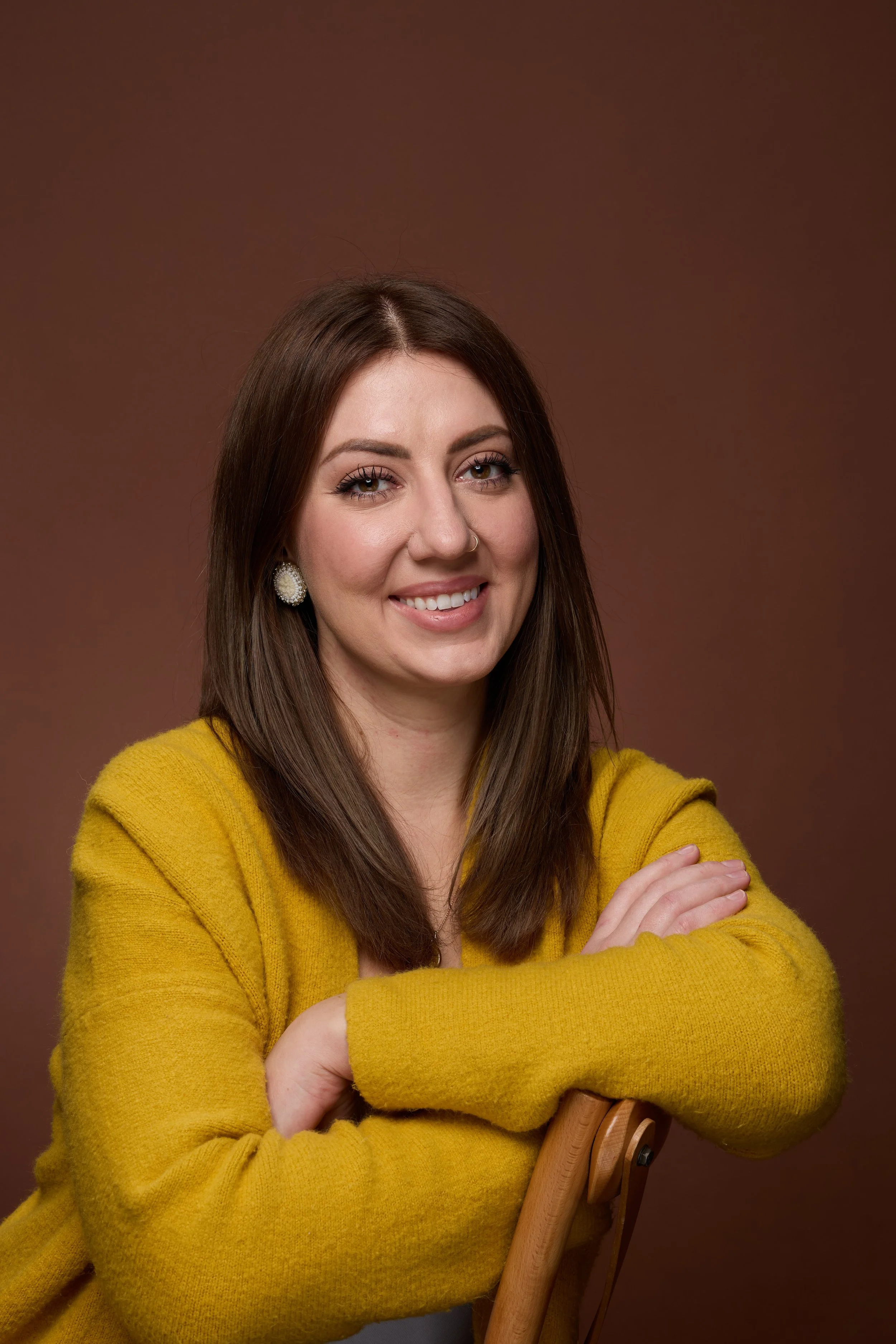 Portrait of a smiling woman with long brown hair, wearing a yellow sweater and earrings, against a brown background.