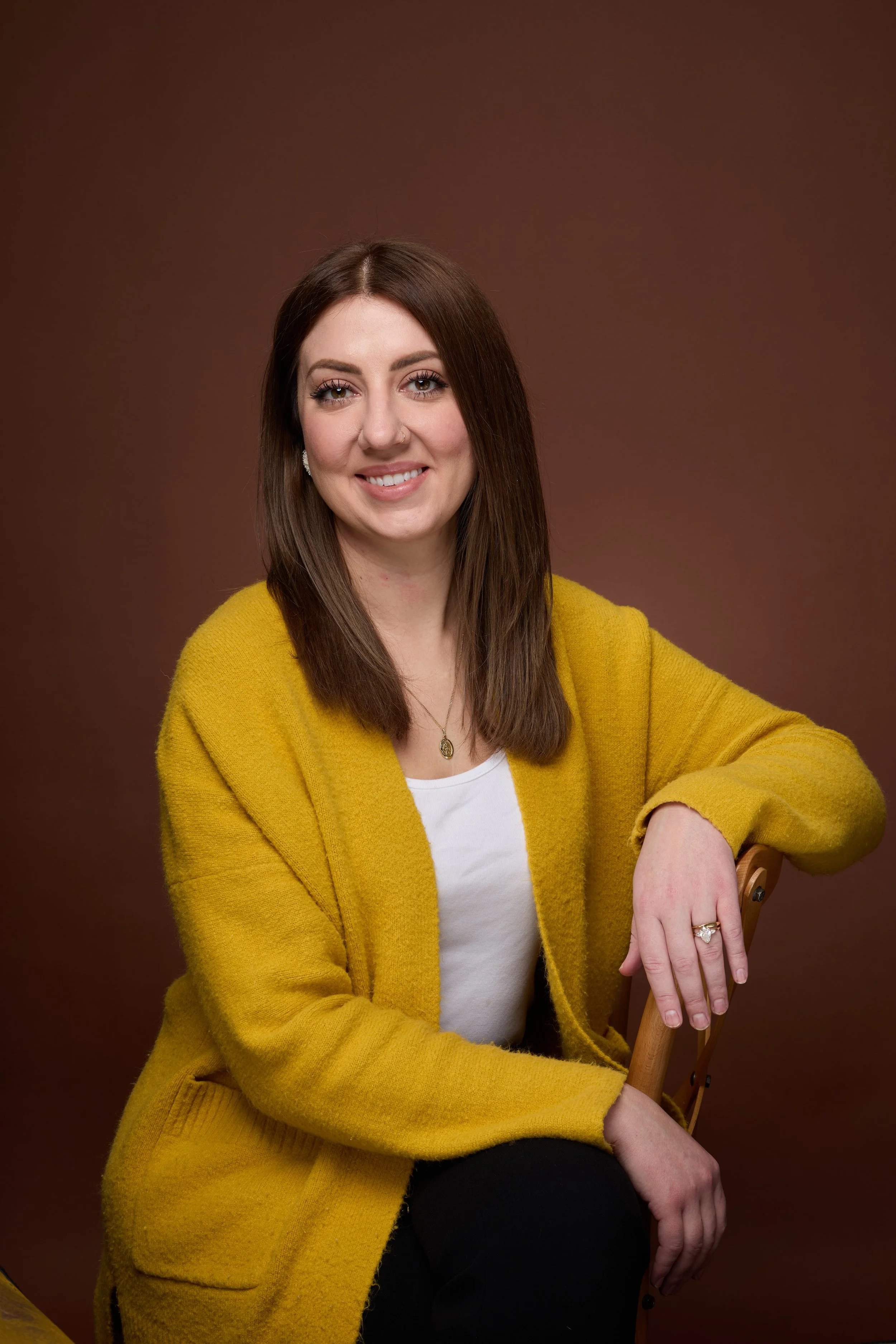 A young woman with brown hair and blue eyes smiling, wearing a yellow cardigan over a white shirt, sitting on a wooden chair against a brown background.