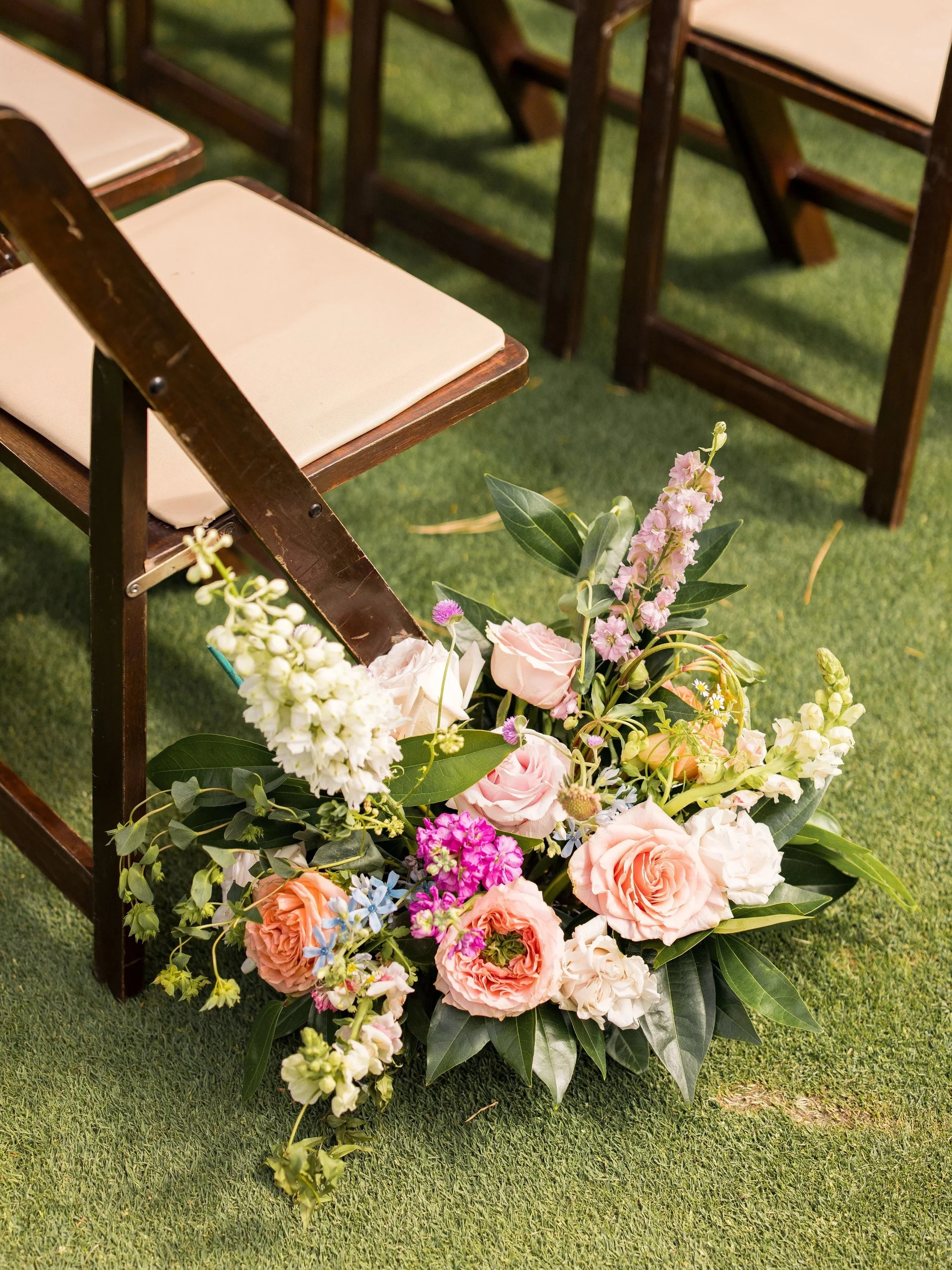 A floral arrangement with pink, white, purple, and peach flowers and green leaves on grass, with wooden chairs nearby.